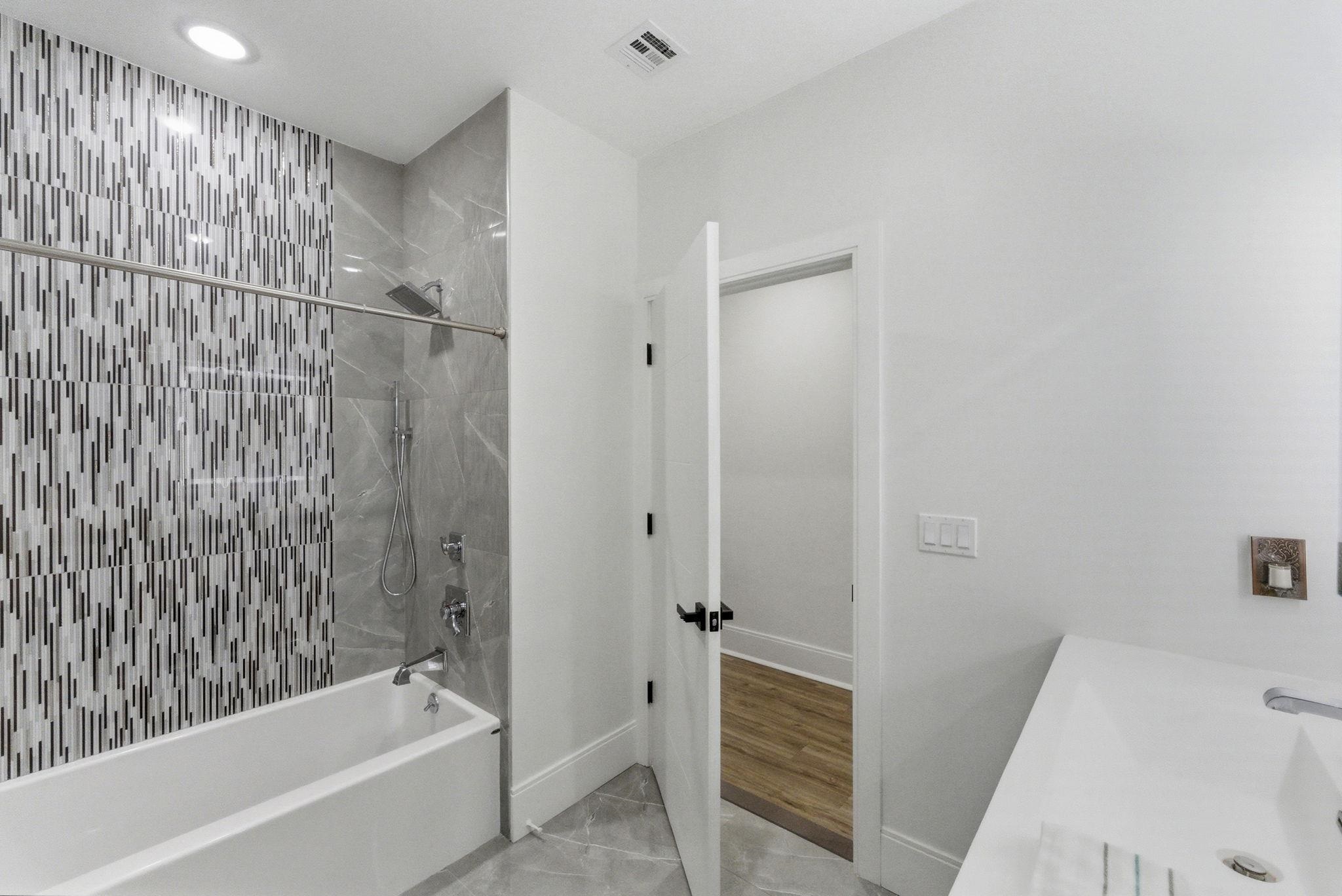 Bathroom with a white tub, gray tile, mosaic accent wall, and open doorway to a wood-floored room.