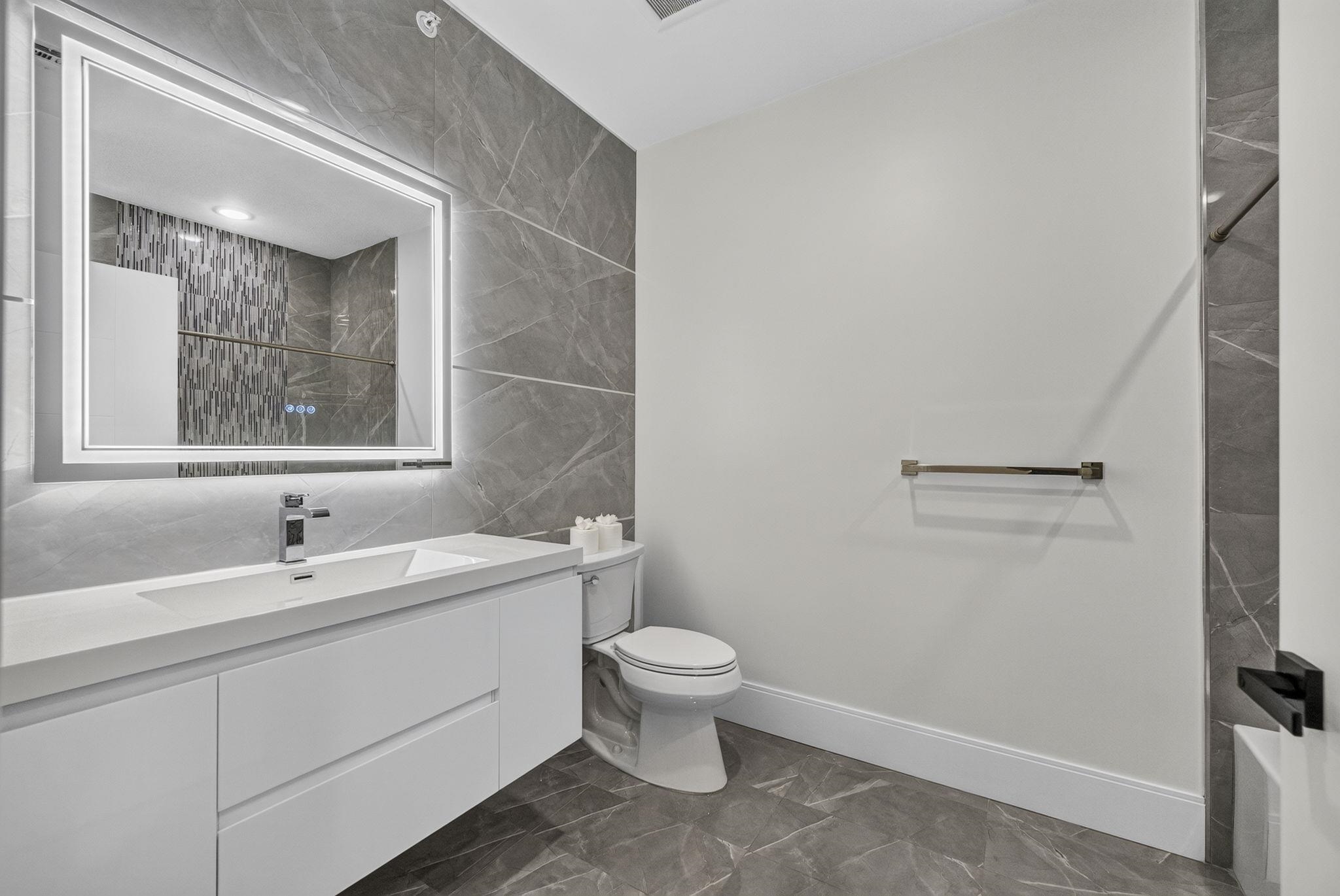 Modern bathroom with a white vanity, illuminated mirror, gray tile, and glass shower.