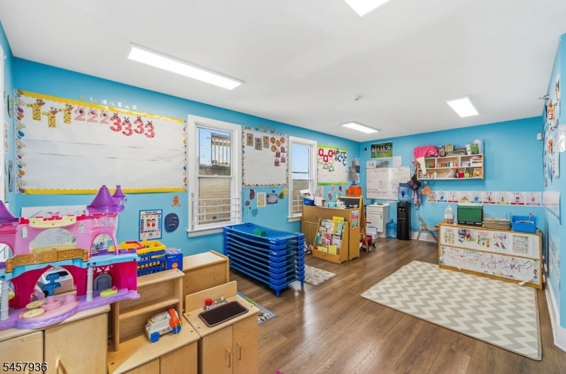 Colorful classroom with toy kitchens, educational displays, stacked blue cots, and a patterned rug.