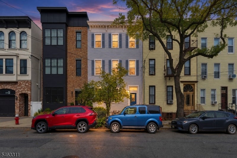 Attached urban homes with varied facades and parked cars beneath a mature tree at dusk.
