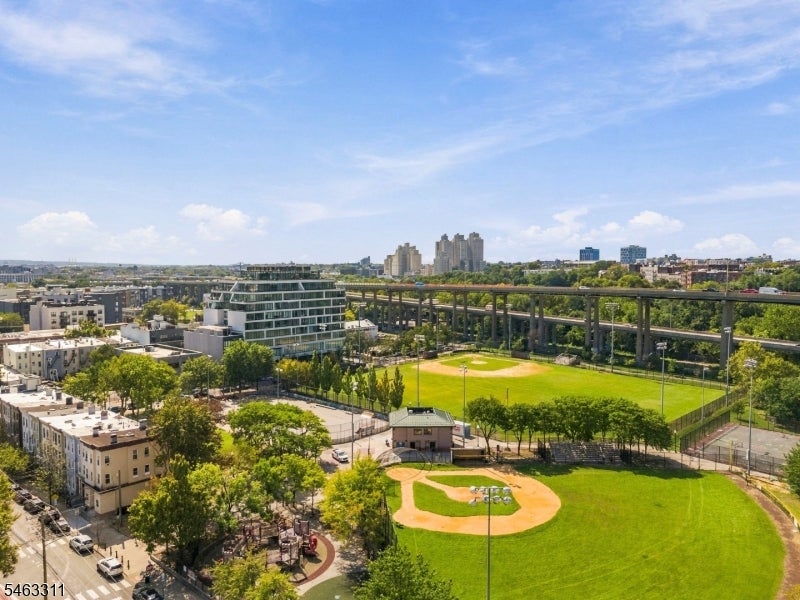 Aerial view over a baseball field, elevated roadway, and Jersey City buildings.