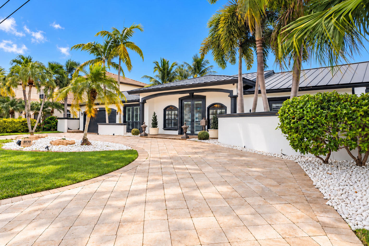 Home exterior with a dark metal roof, arched windows, tiled driveway, and palm-lined landscaping.