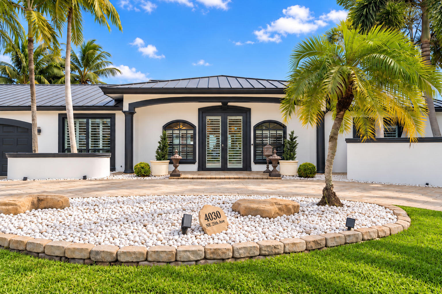 White single-story home with dark trim and arched windows, framed by palms above a white-stone front garden.