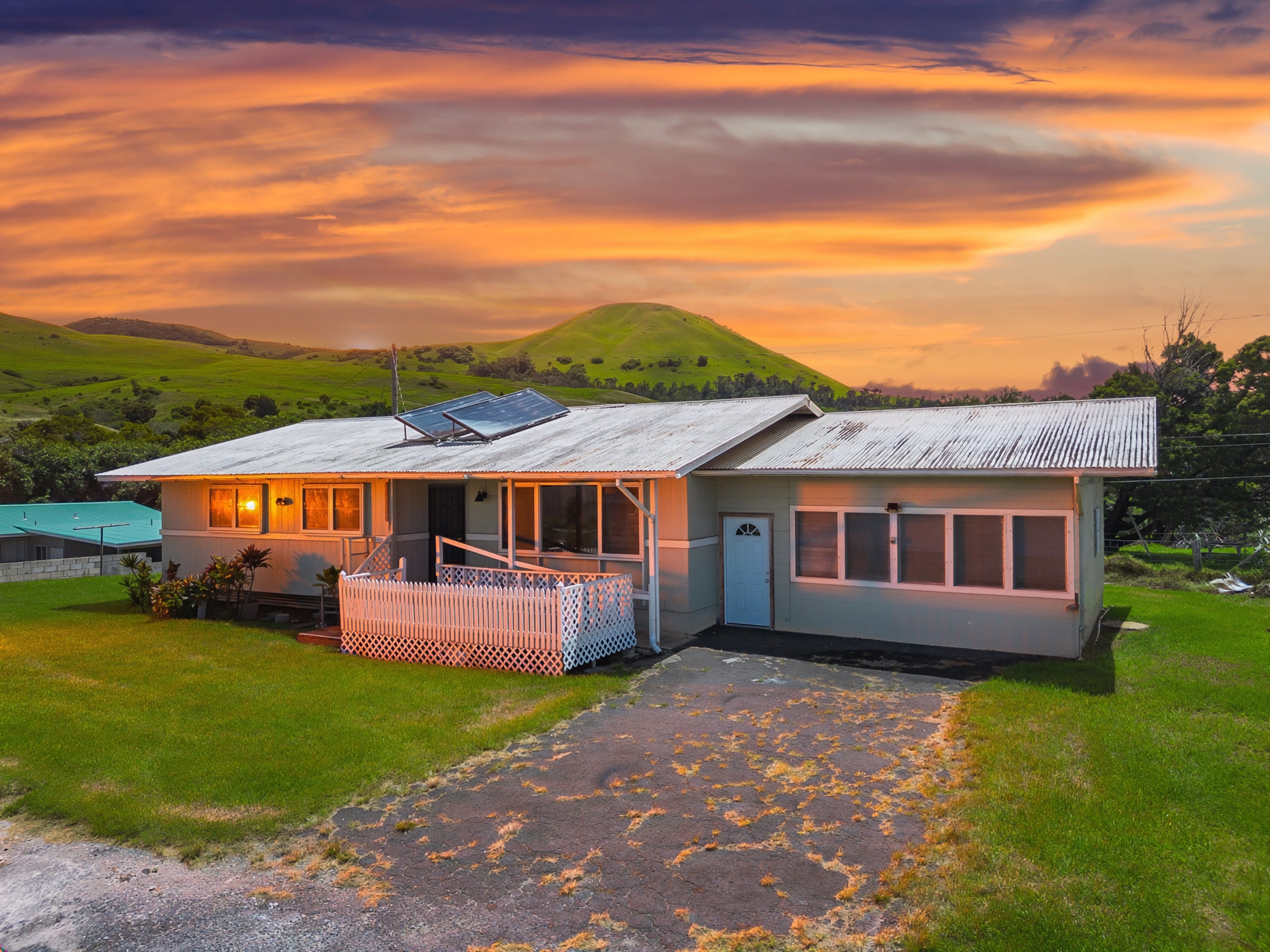 Single-story home with rooftop solar panels, a fenced porch, and green hills at sunset.