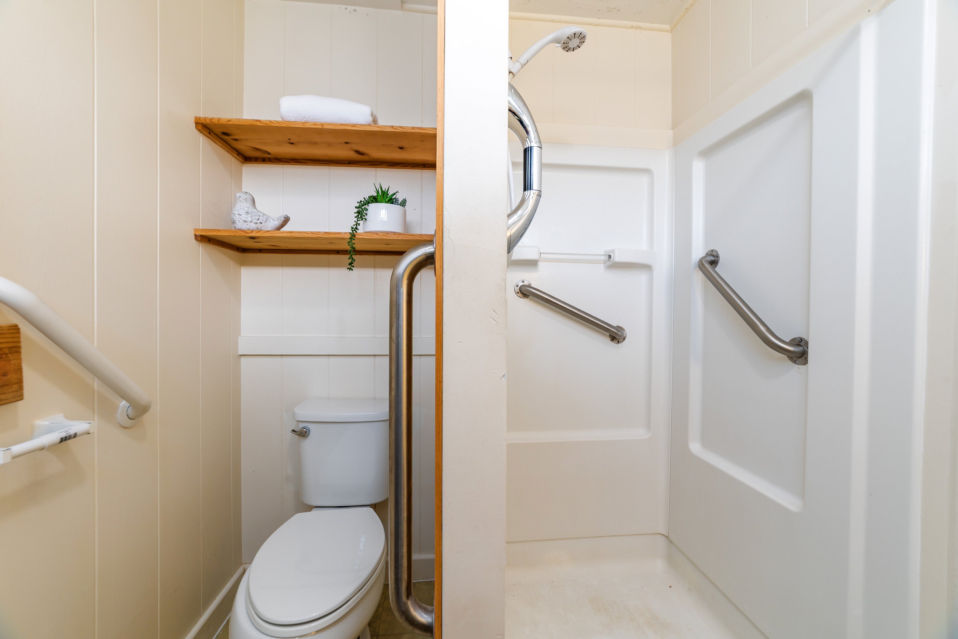 Bathroom with an accessible shower, stainless steel grab bars, and wooden shelves above the toilet.