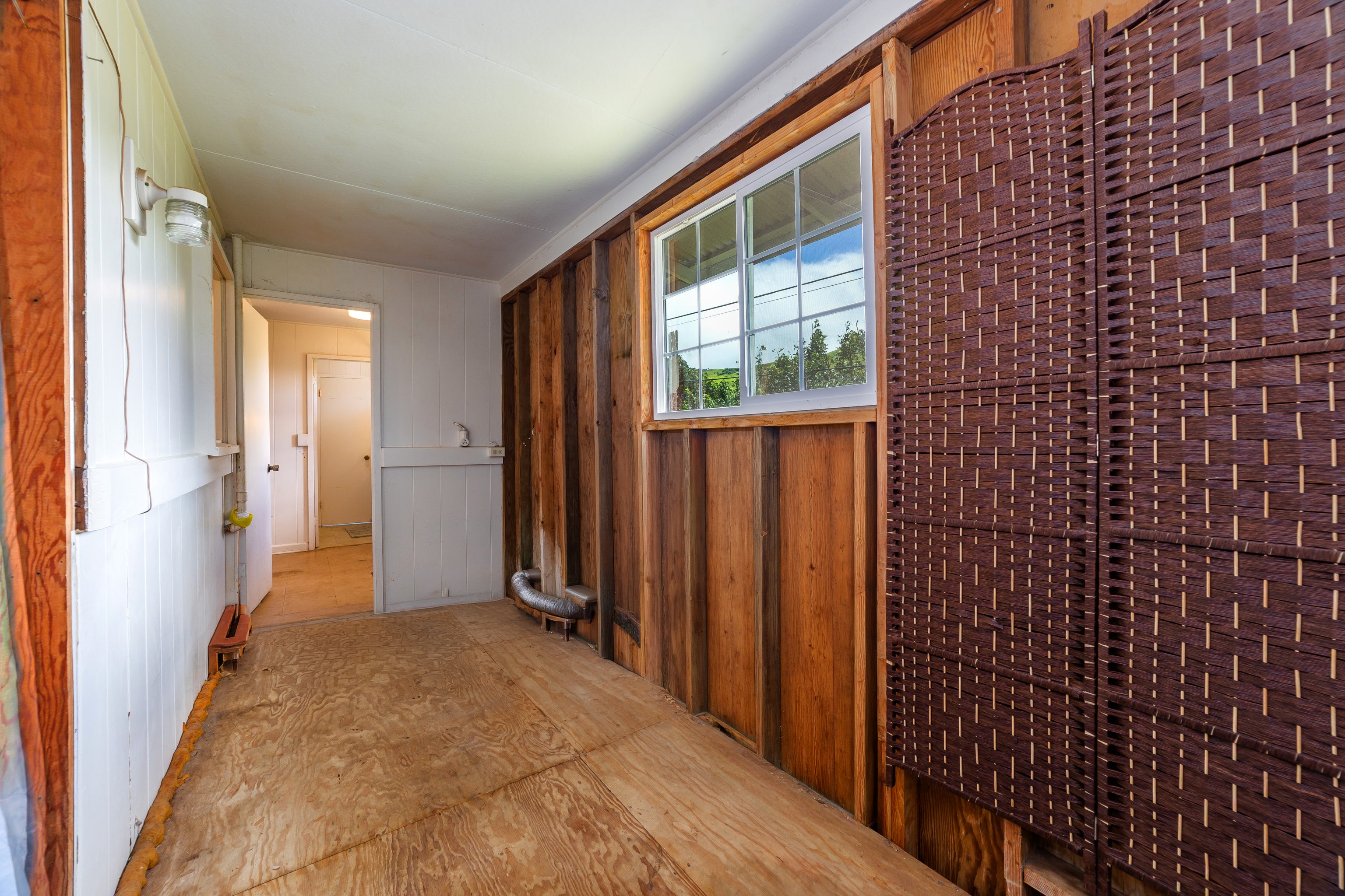 Unfinished interior room with plywood flooring, wood-paneled walls, a window, and a woven wall covering.