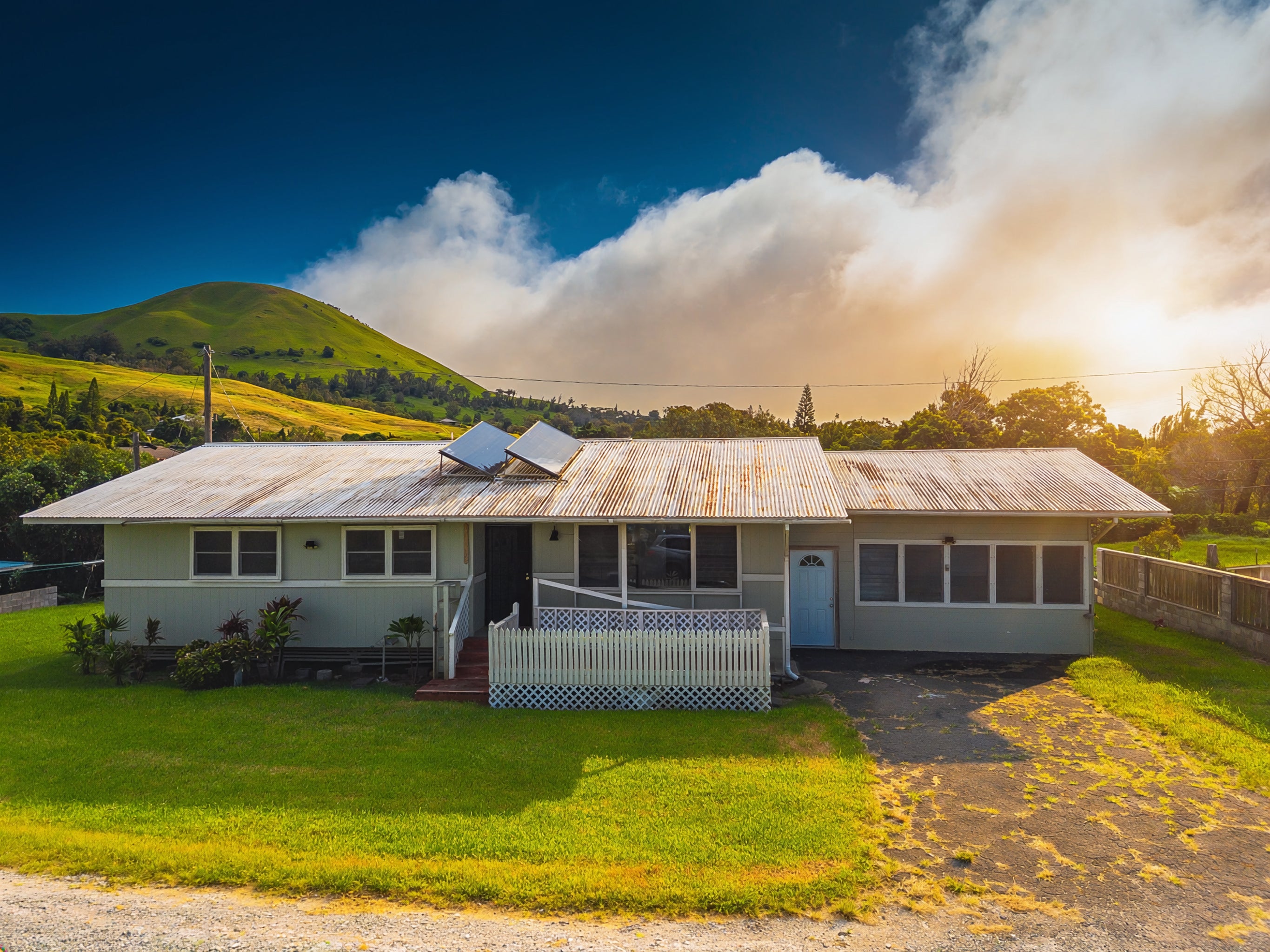 Single-story home with solar panels on a corrugated roof, green lawn, and hillside behind it.