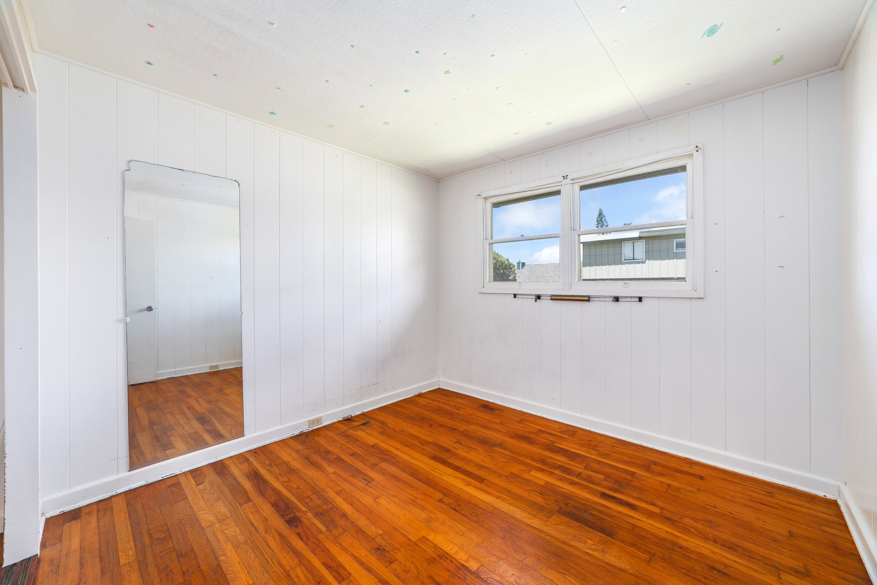 Empty bedroom with reddish-brown wood floors, a mirrored closet door, and a window overlooking a neighboring building.