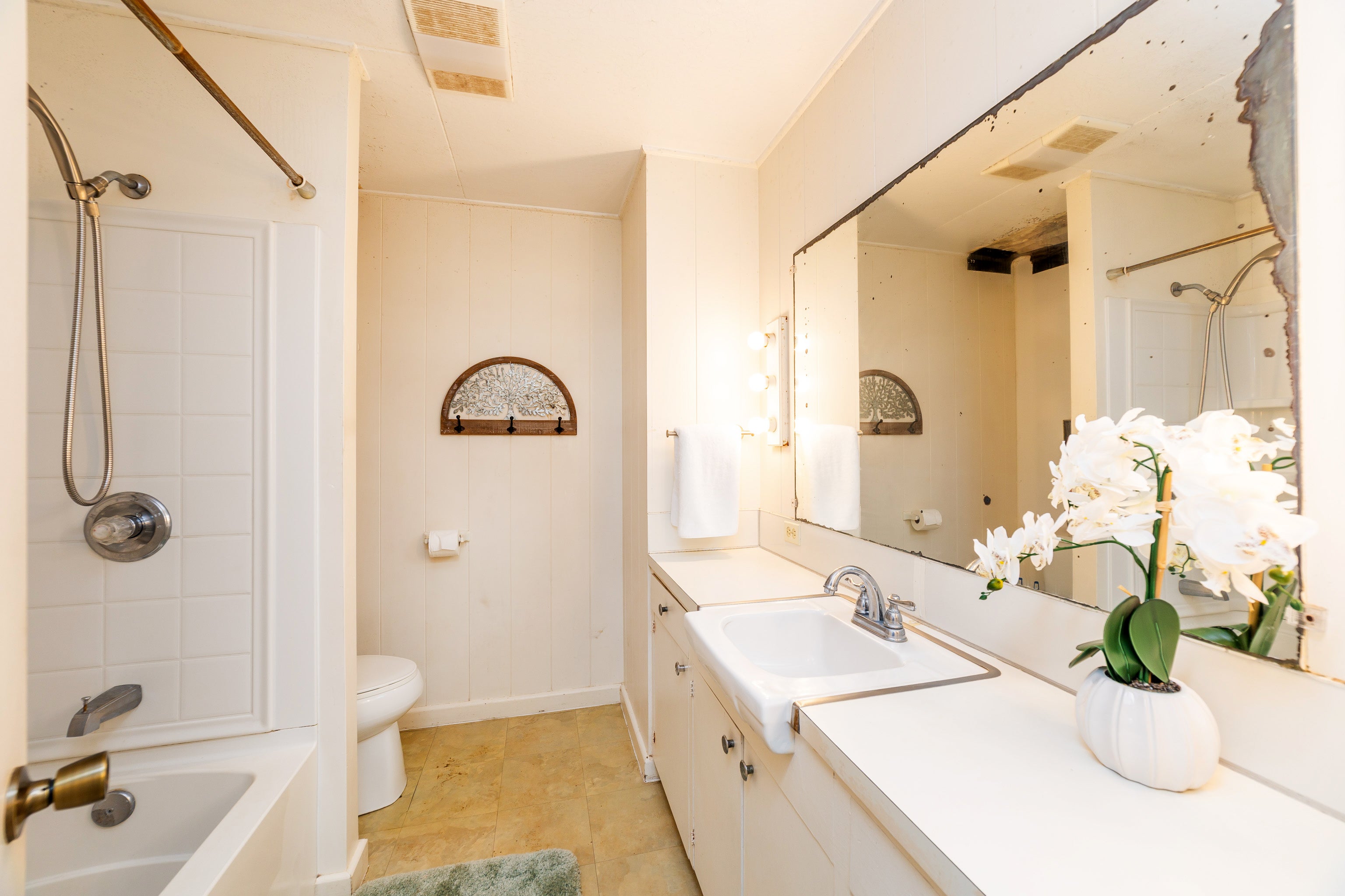 Bathroom with a tiled tub and shower, long white vanity, and a white orchid beside a worn mirror.