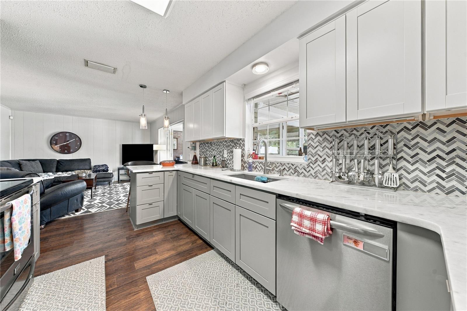 Open-plan kitchen with white cabinets, gray herringbone backsplash, marble-look counters, and a dark sectional.