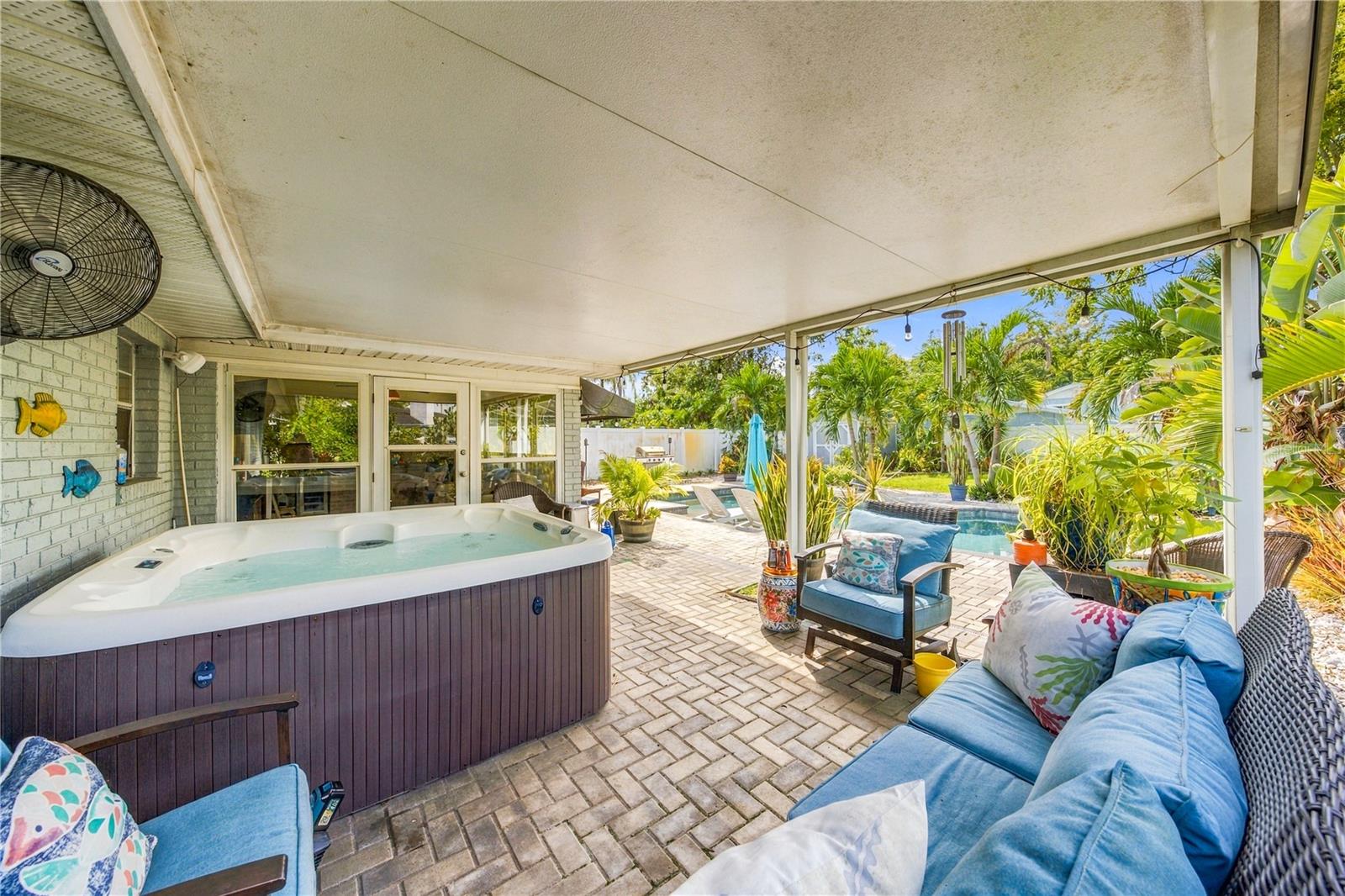 Covered patio with a hot tub, cushioned seating, and a pool amid tropical landscaping.