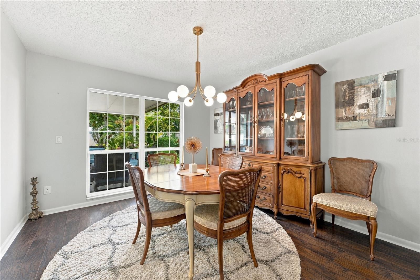 Dining room with a wooden table, glass-front china cabinet, and window overlooking tropical greenery.