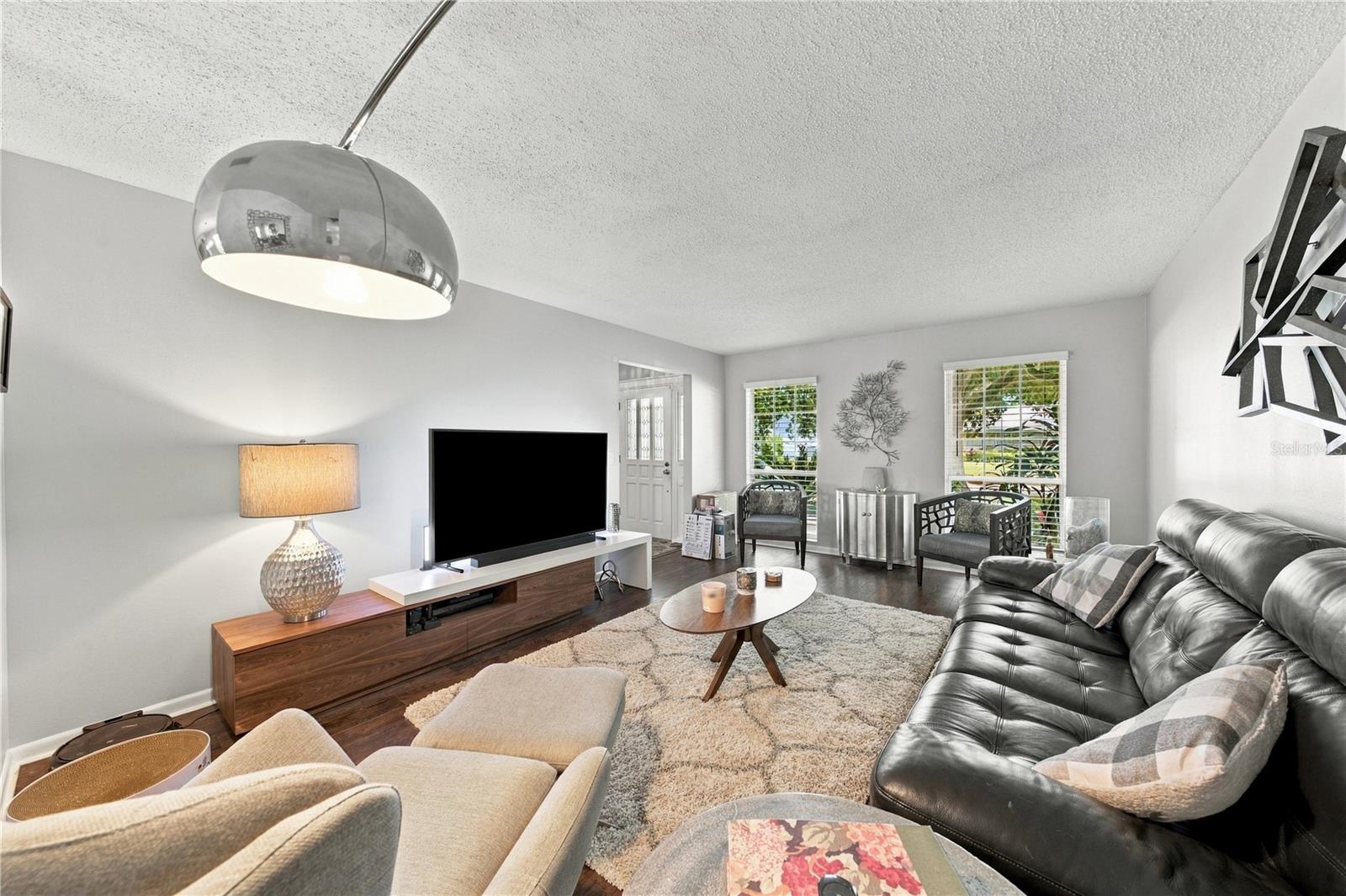 Furnished living room with a black leather sofa, cream chairs, and a television beside two windows.