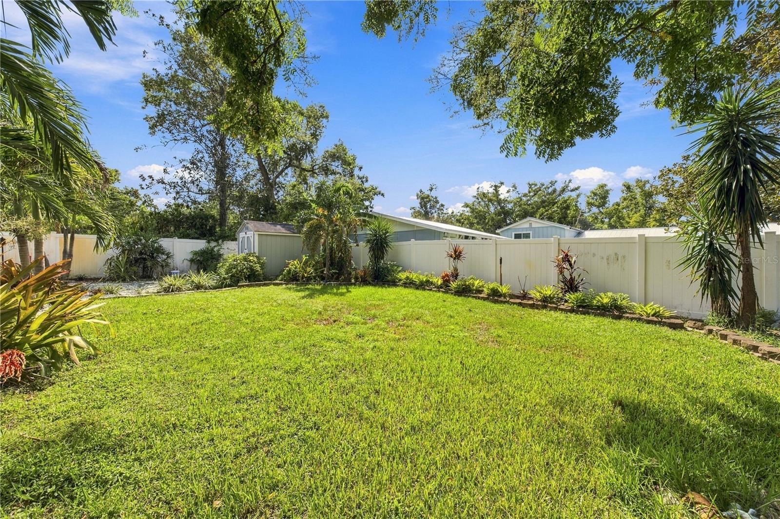 Spacious backyard with a green lawn, white privacy fence, and mature tropical landscaping.