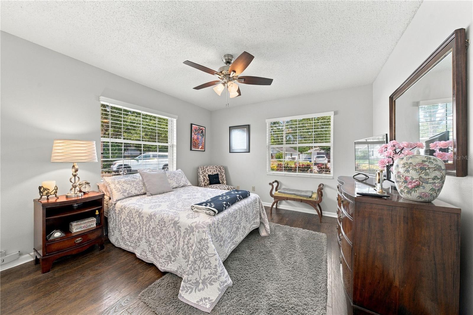 Furnished bedroom with a floral bedspread, dark wood furniture, and two bright windows.