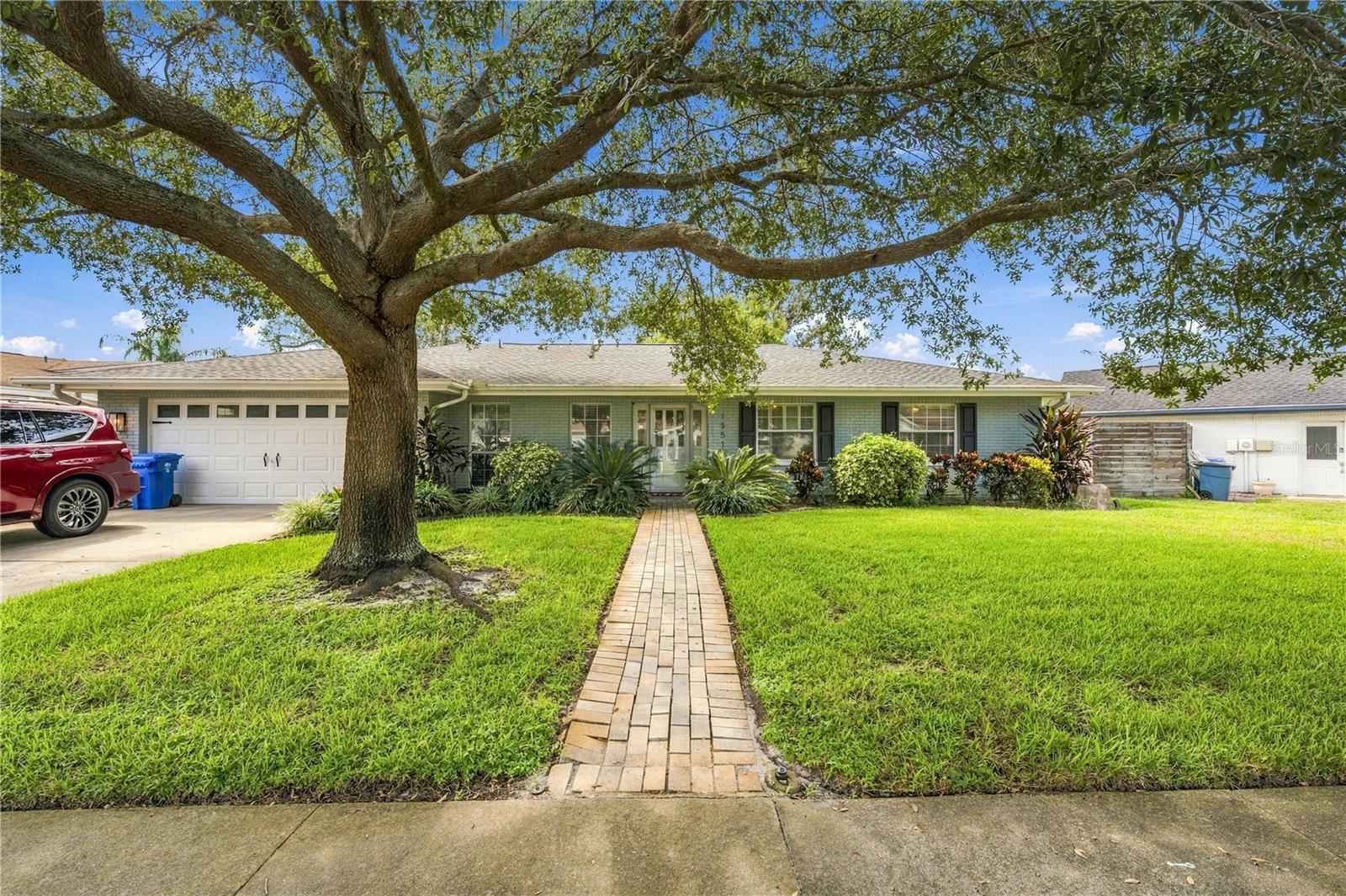 Single-story blue home with an attached garage, brick walkway, and large oak tree in the front yard.