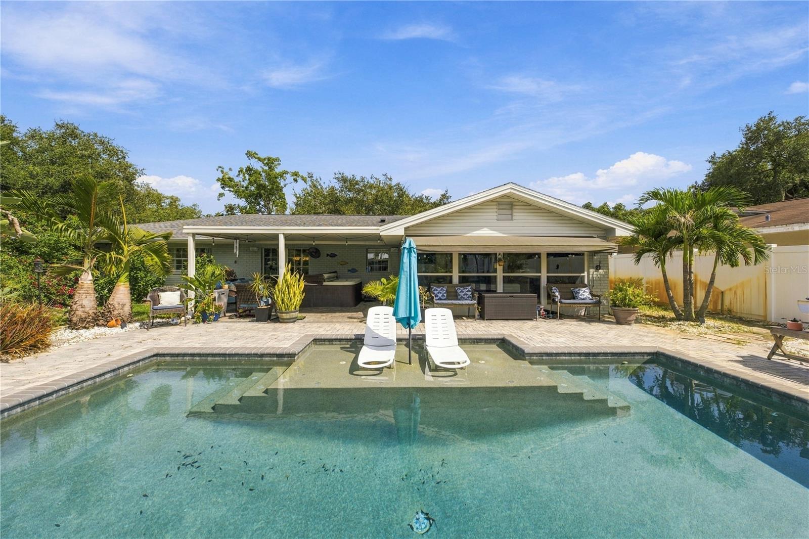 Rear of a single-story home with a pool, patio seating, and tropical landscaping.