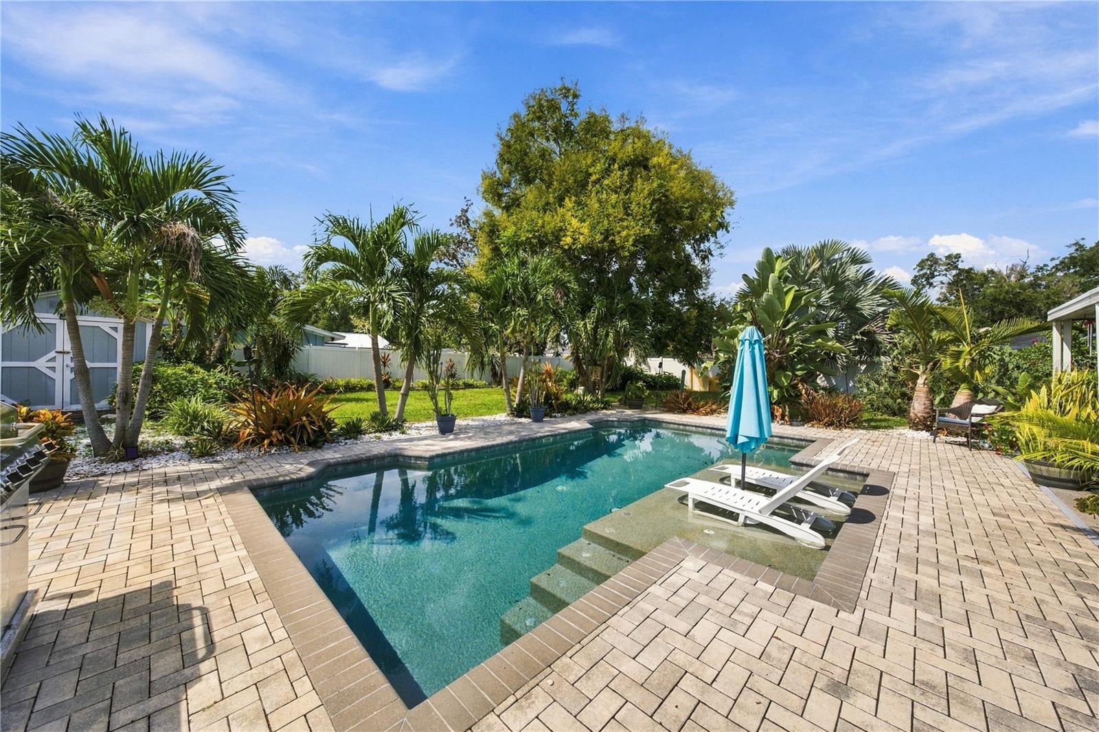 Backyard pool with tropical landscaping, paved patio, and white lounge chairs.