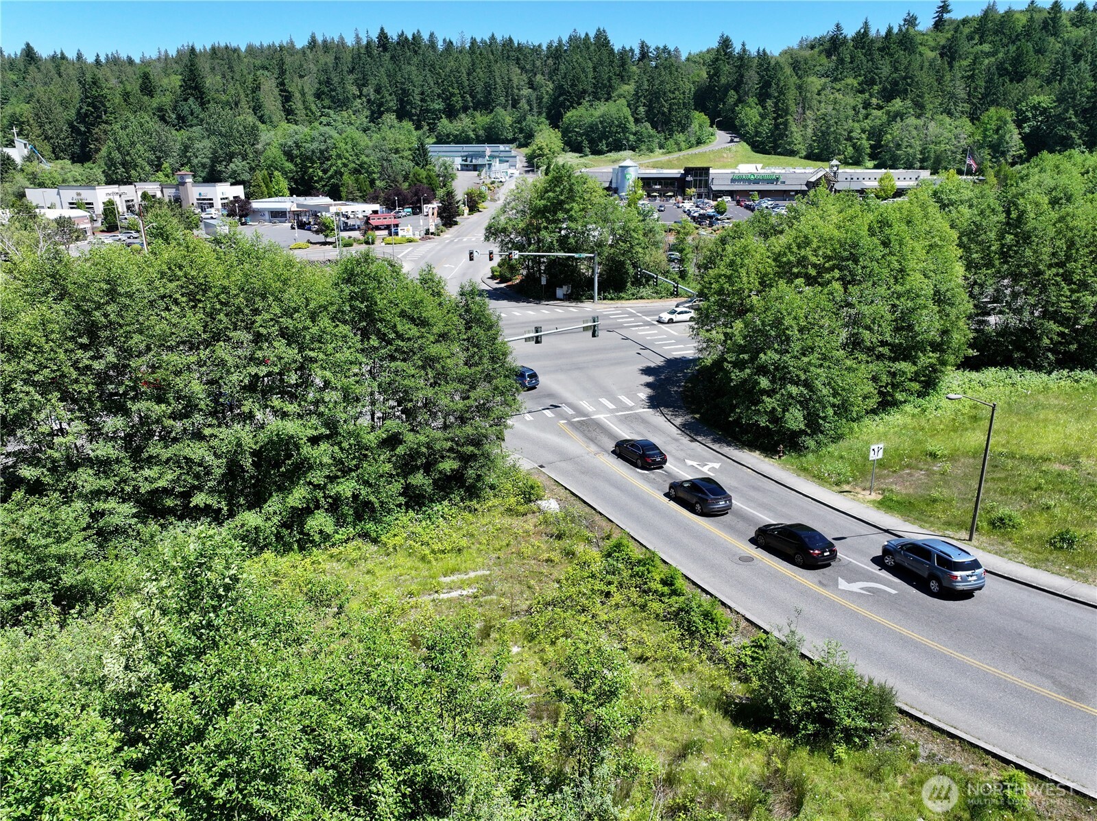 Aerial view of vacant brush-covered land beside a busy intersection, with nearby shops and evergreen forest.