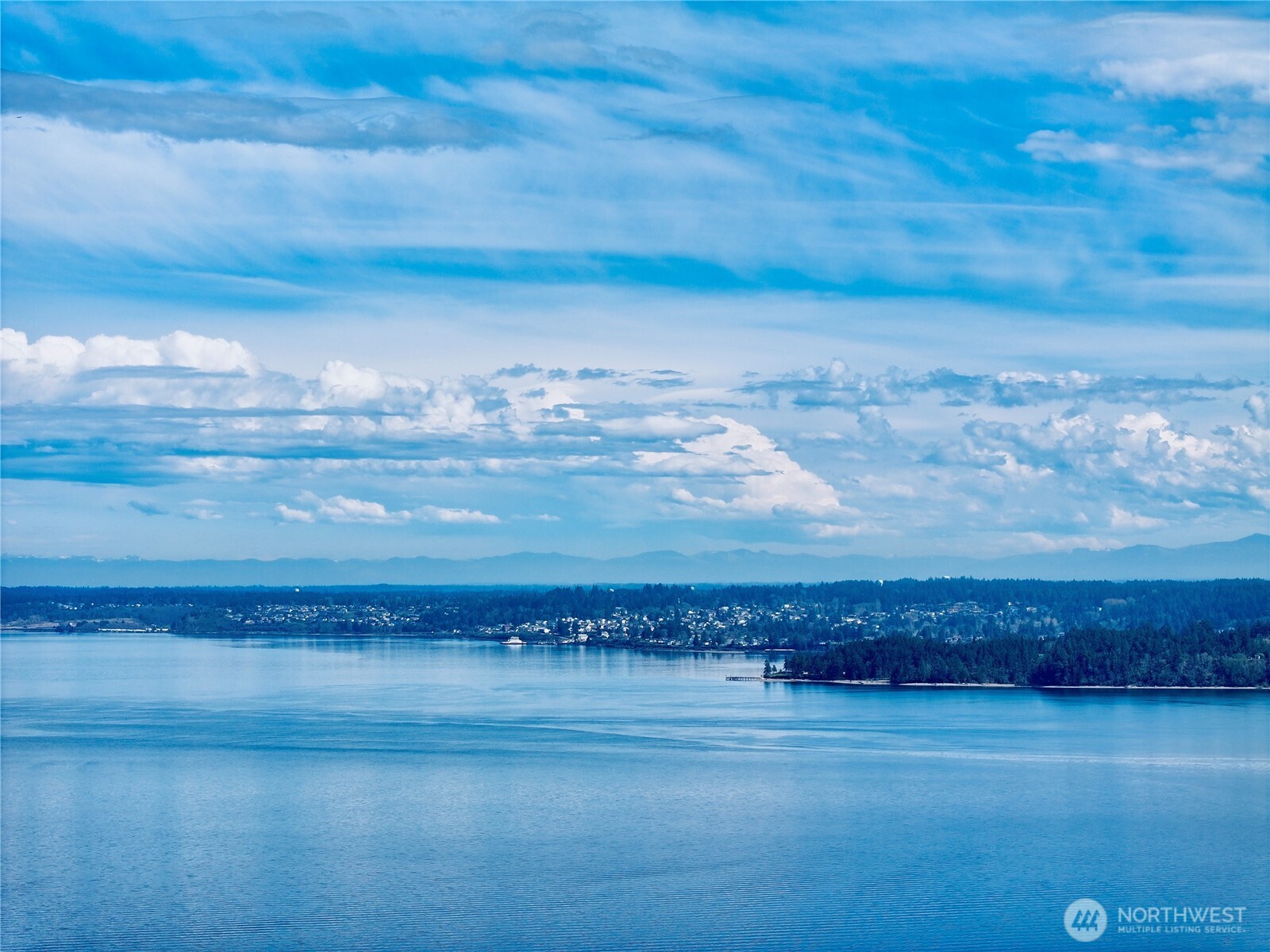 Waterfront panorama with a wooded shoreline, distant homes, and mountains beneath broad clouds.