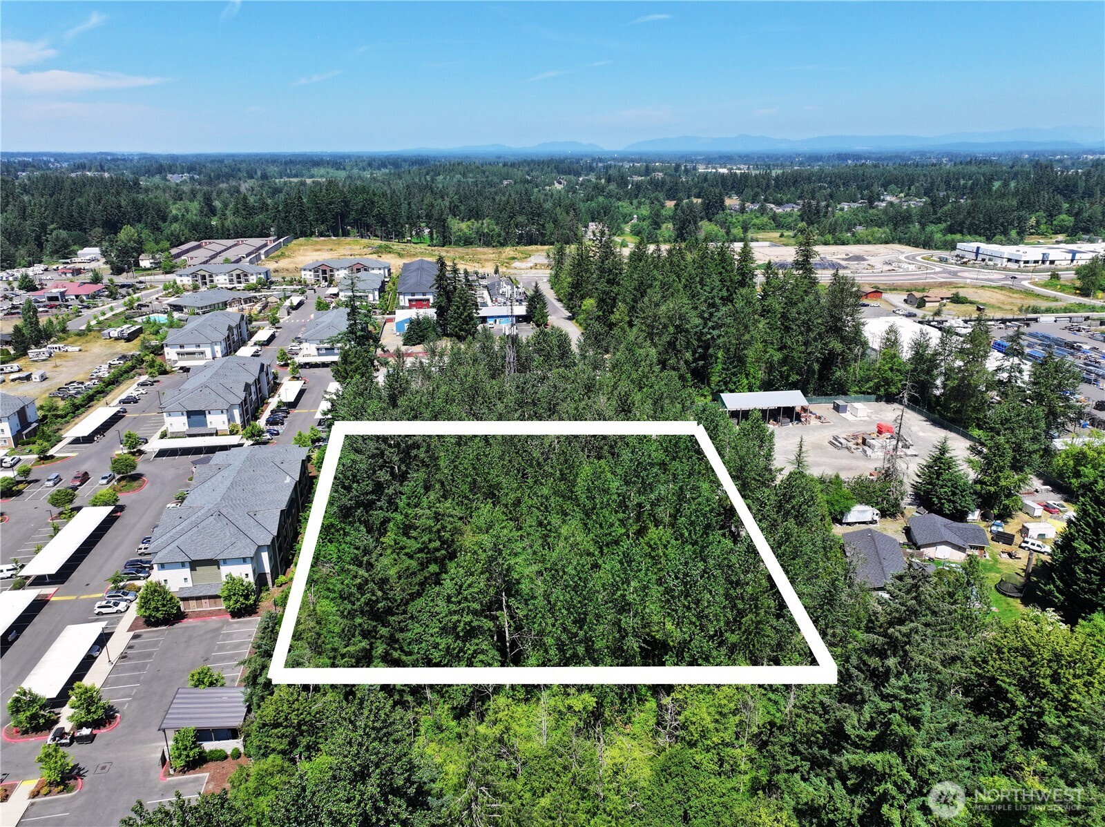 Aerial view of a wooded parcel outlined in white, with nearby buildings and distant mountains.
