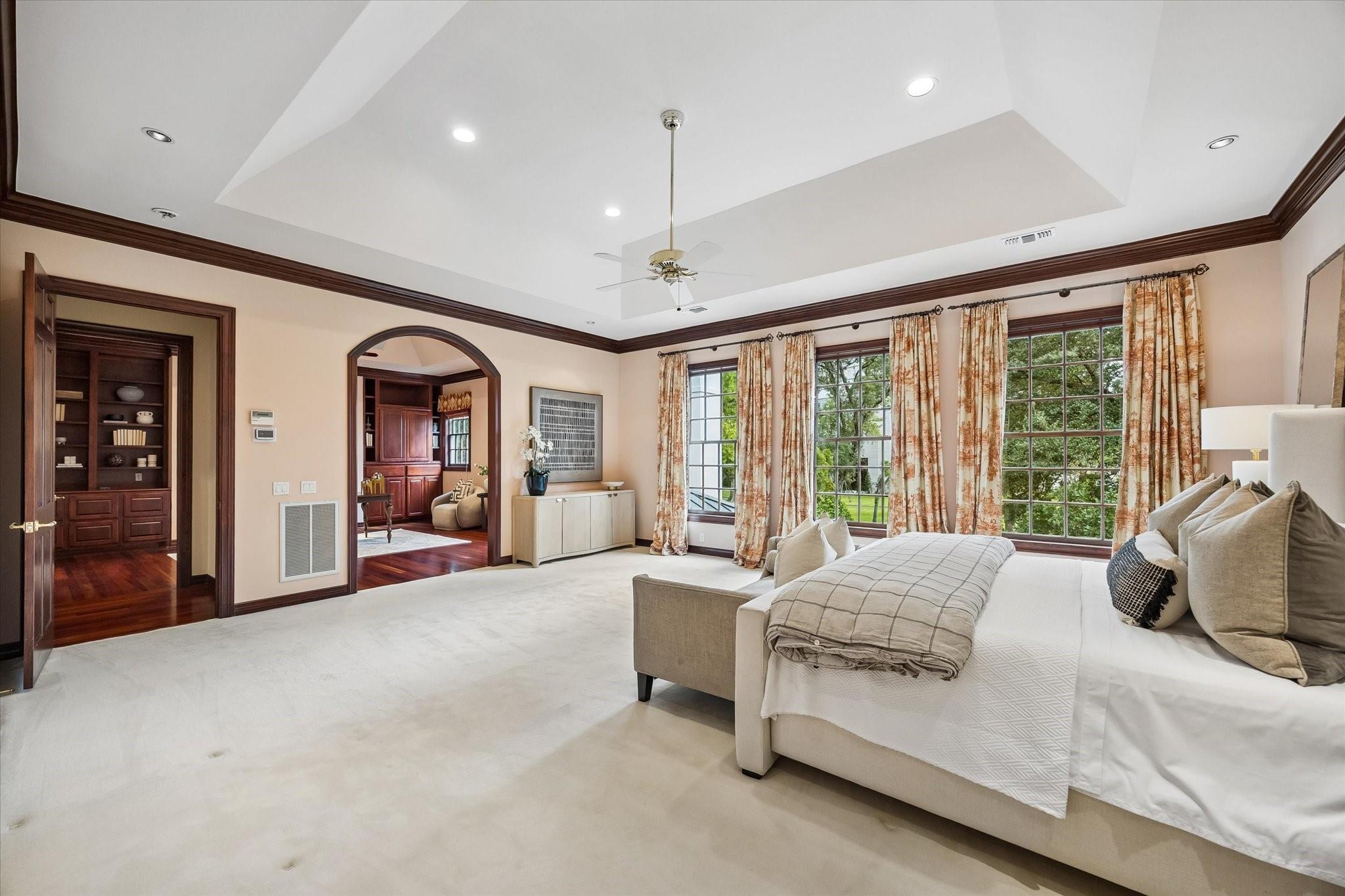 Spacious bedroom with a white bed, tall curtained windows, tray ceiling, and dark wood trim.