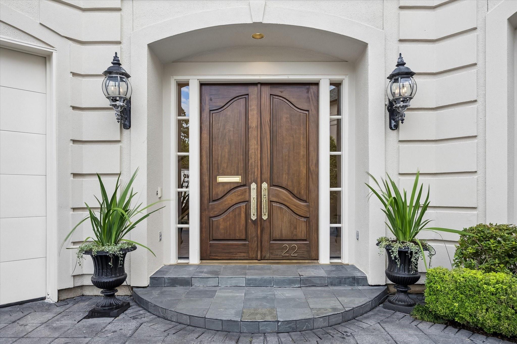 Wooden double entry doors beneath an arch, flanked by lanterns and potted plants.