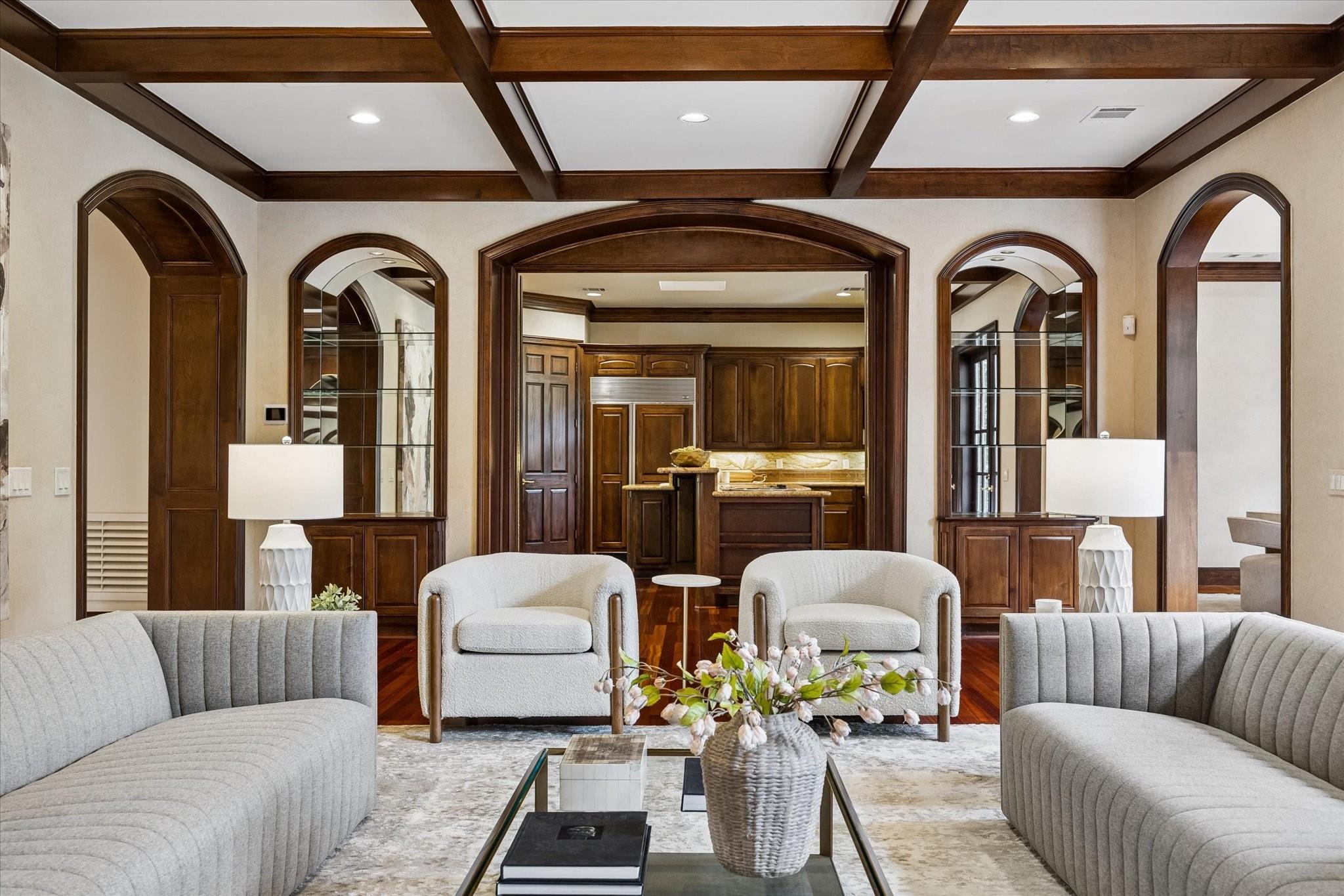 Living room with curved gray sofas, wood ceiling beams, and an arched opening to the kitchen.