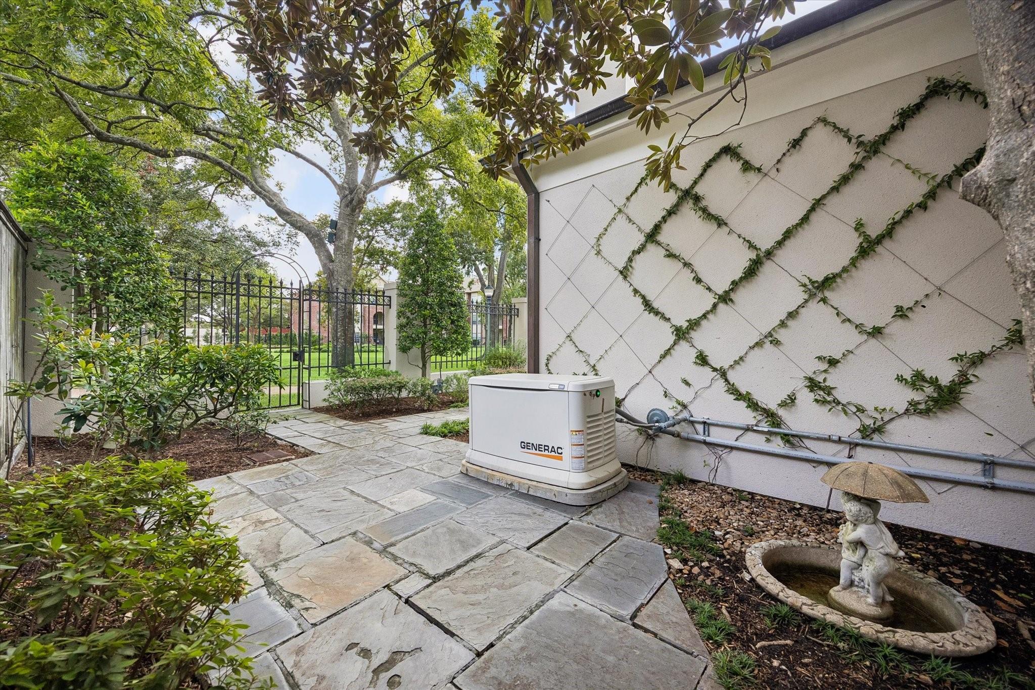 Stone patio with a standby generator, ivy-covered wall, iron gate, and small garden fountain.
