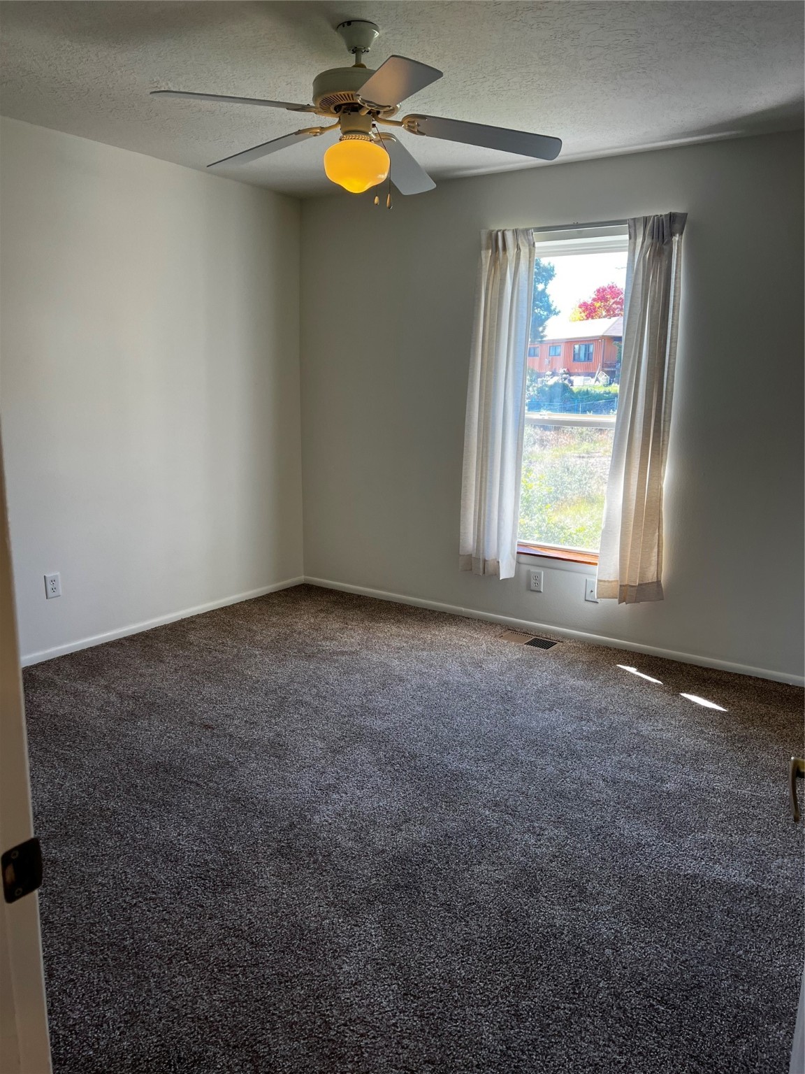 Empty carpeted bedroom with a ceiling fan and curtained window overlooking a neighboring house.