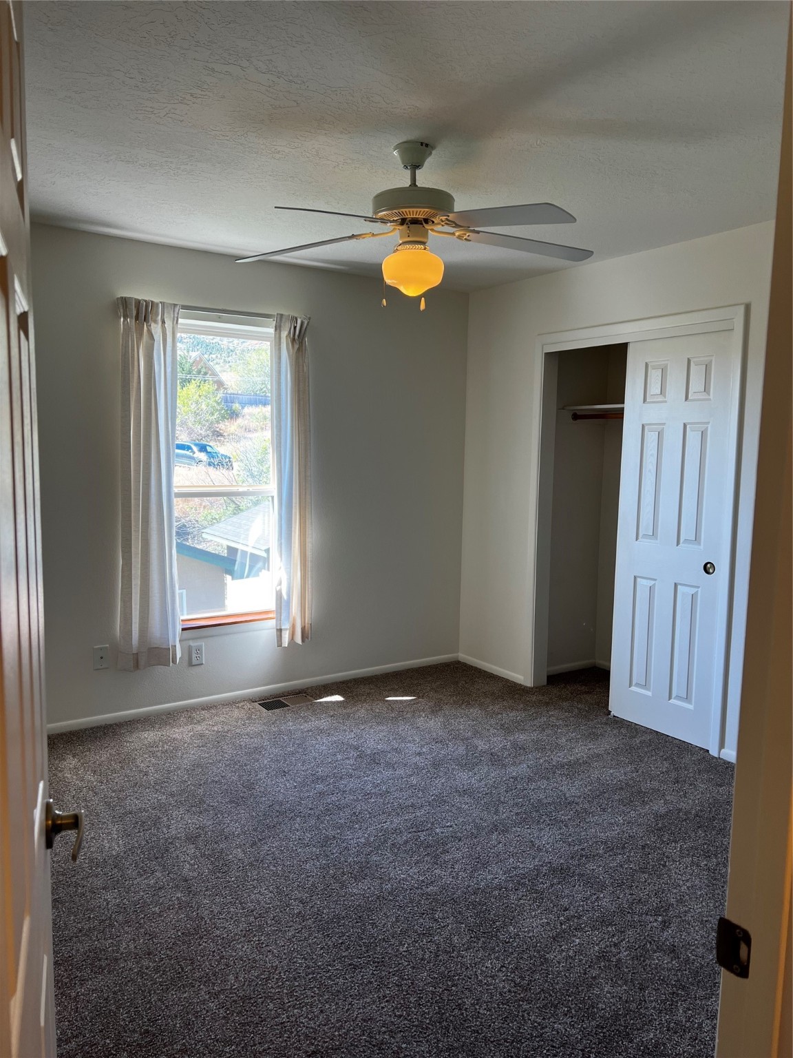 Empty bedroom with gray carpet, a curtained window, ceiling fan, and open closet.