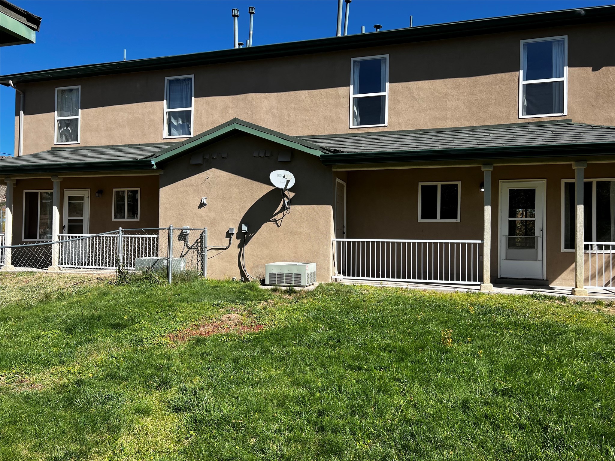 Two-story tan stucco home with green roof trim, covered porch, fenced patio, and grassy lawn.