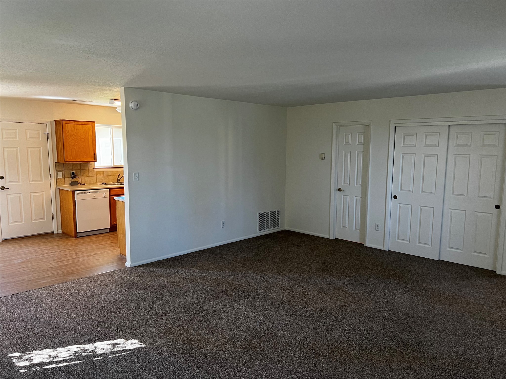 Unfurnished living room with dark carpet opening onto a kitchen with wood cabinets.