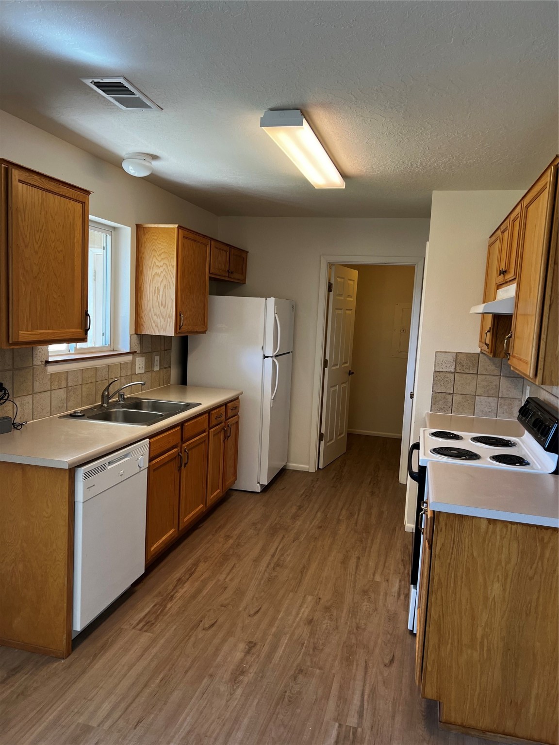 Galley kitchen with wood cabinets, a window over the sink, white appliances, and wood-look flooring.
