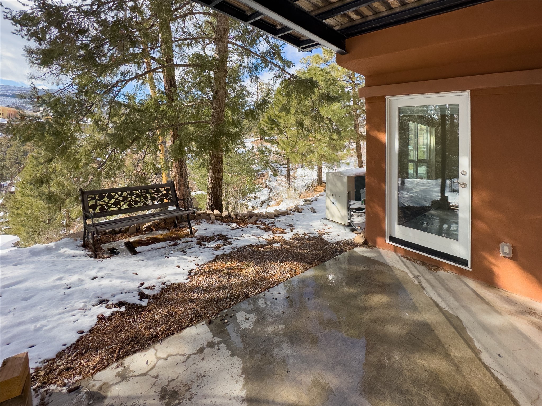 Covered concrete patio with a glass door, bench, and snow-dusted pines.