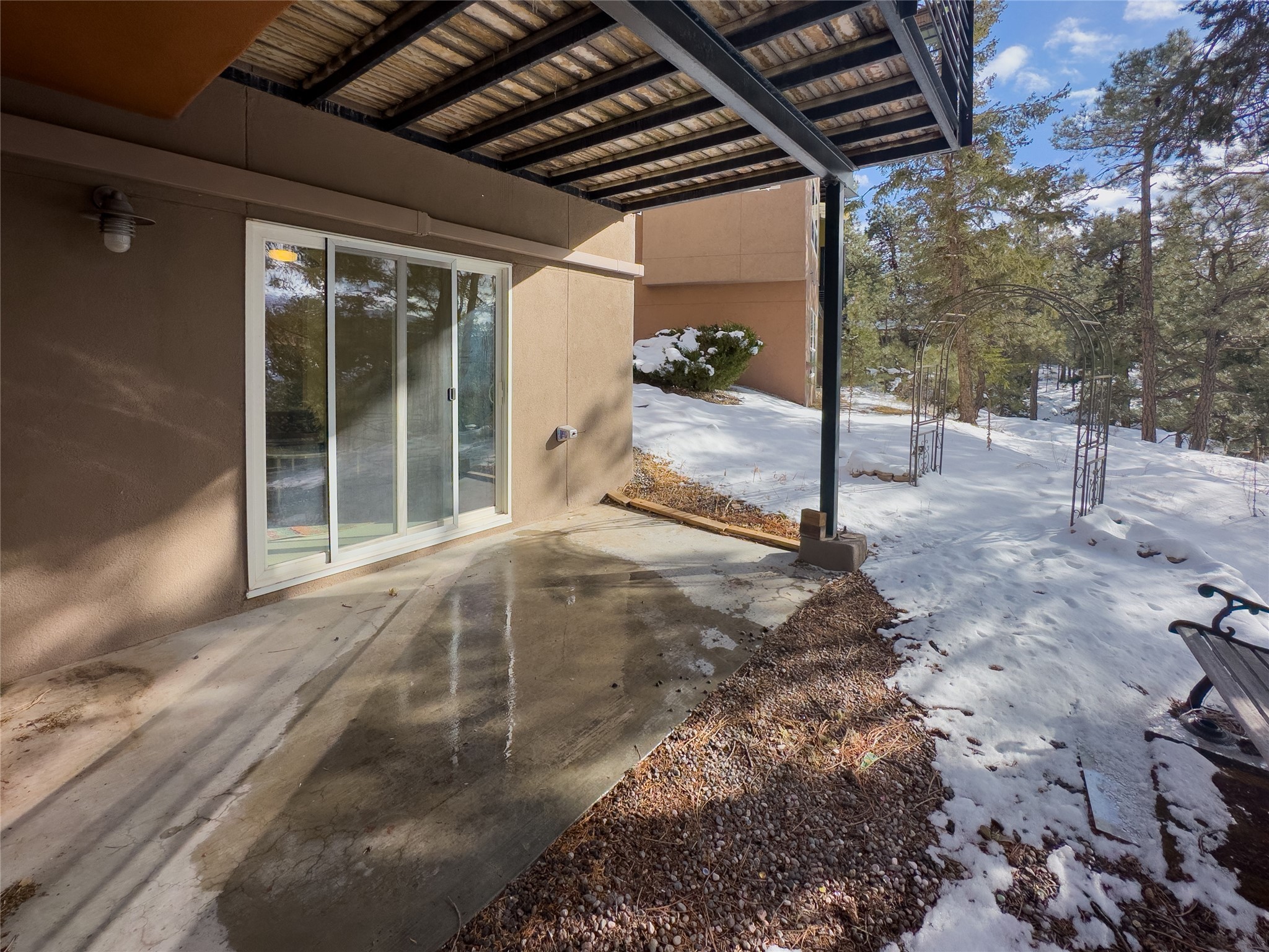Covered concrete patio beside a tan home, opening onto a snowy yard with pine trees and a metal arbor.