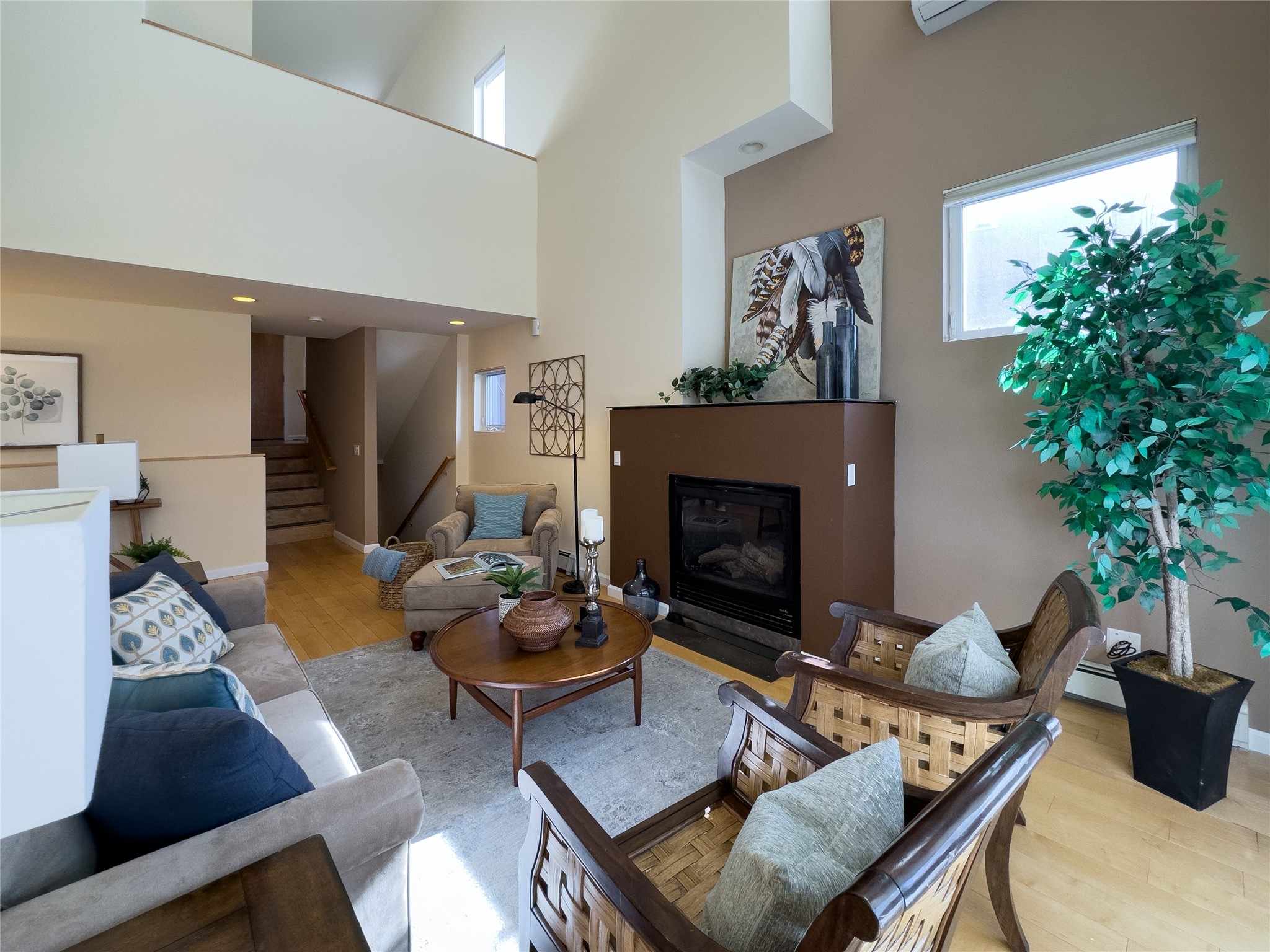 Vaulted living room with a fireplace, woven armchairs, tall windows, and stairs to an upper level.