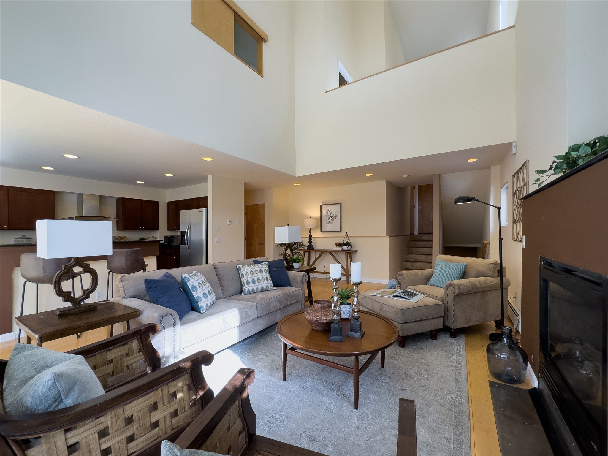 Open-plan living room with a double-height ceiling, staged seating, fireplace, and kitchen beyond.