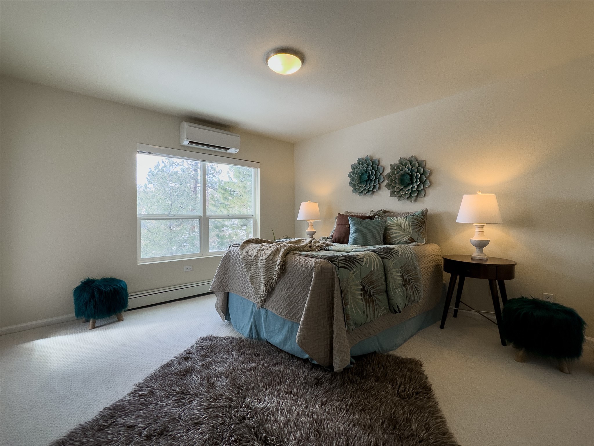 Staged bedroom with muted green bedding, bedside lamps, a large window, and plush rugs.