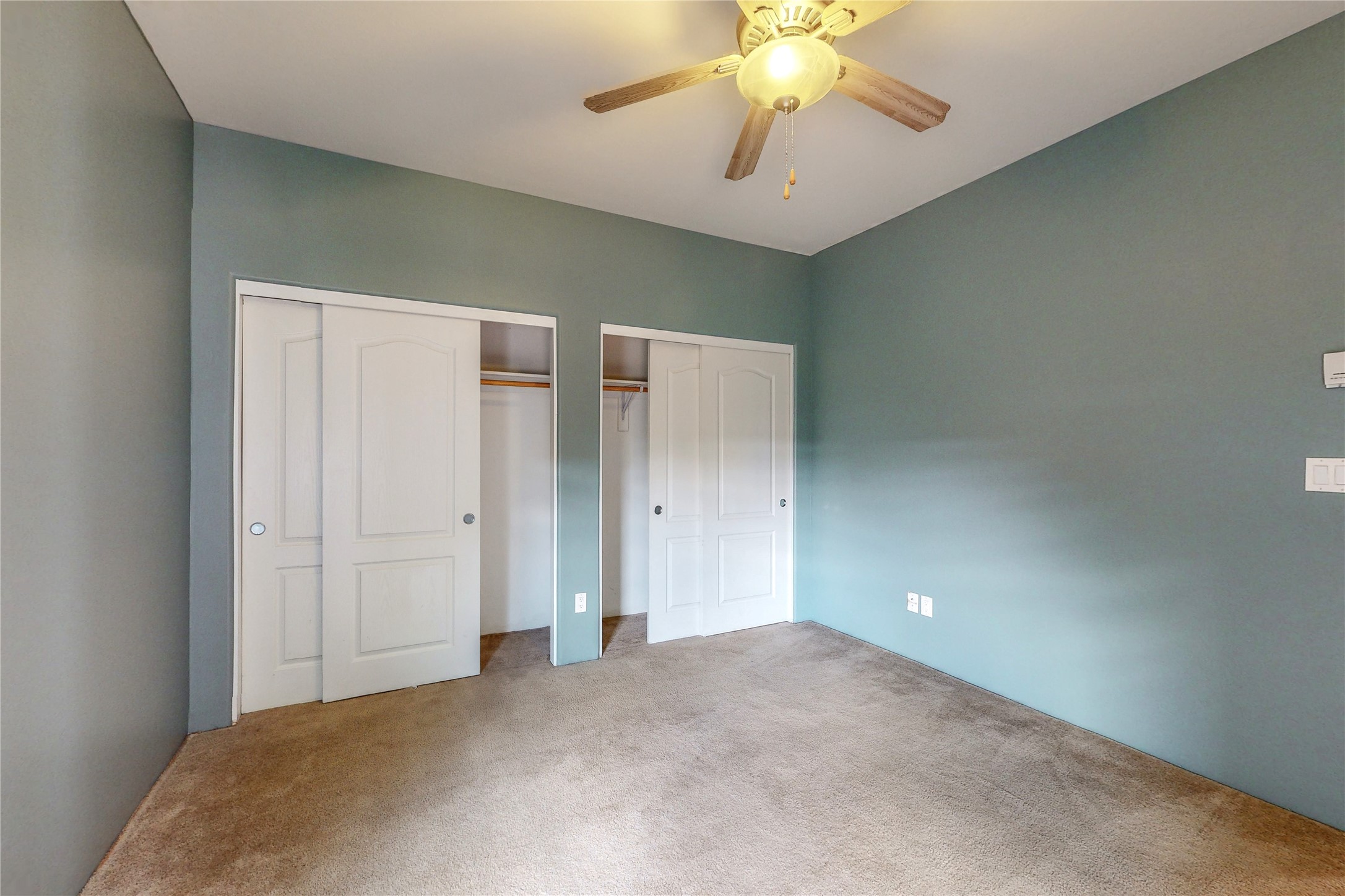 Empty bedroom with two closets, beige carpet, and a lit ceiling fan.