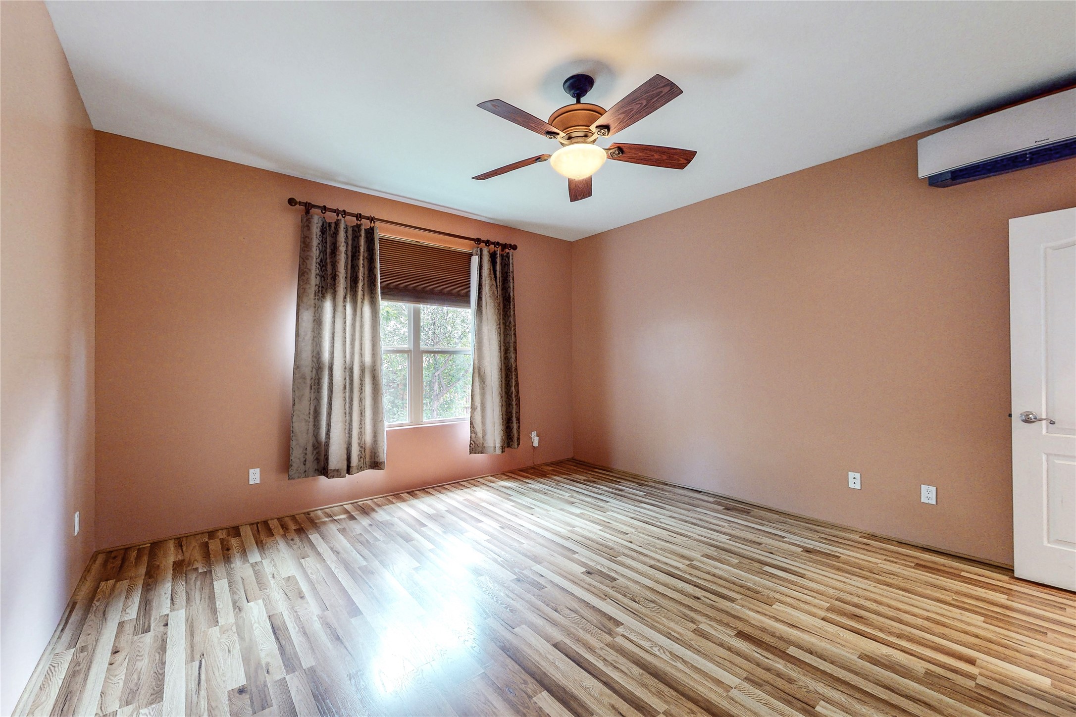 Empty bedroom with wood-look flooring, a curtained window, ceiling fan, and wall-mounted air conditioner.