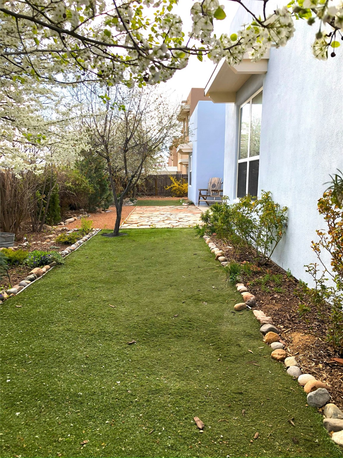 Side yard with a stone-edged lawn, flowering trees, and a flagstone patio beside a stucco home.