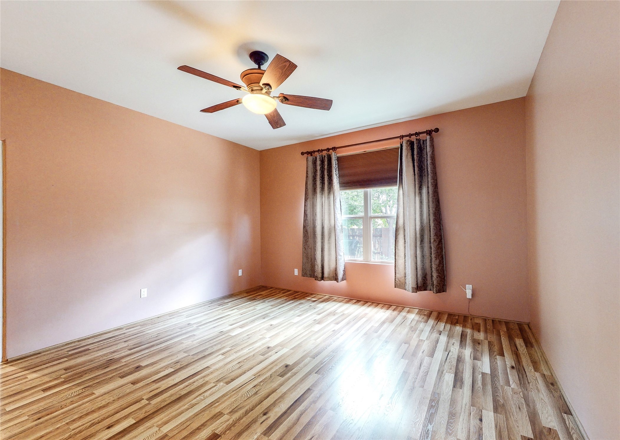 Empty bedroom with wood-look flooring, peach walls, curtained window, and ceiling fan.