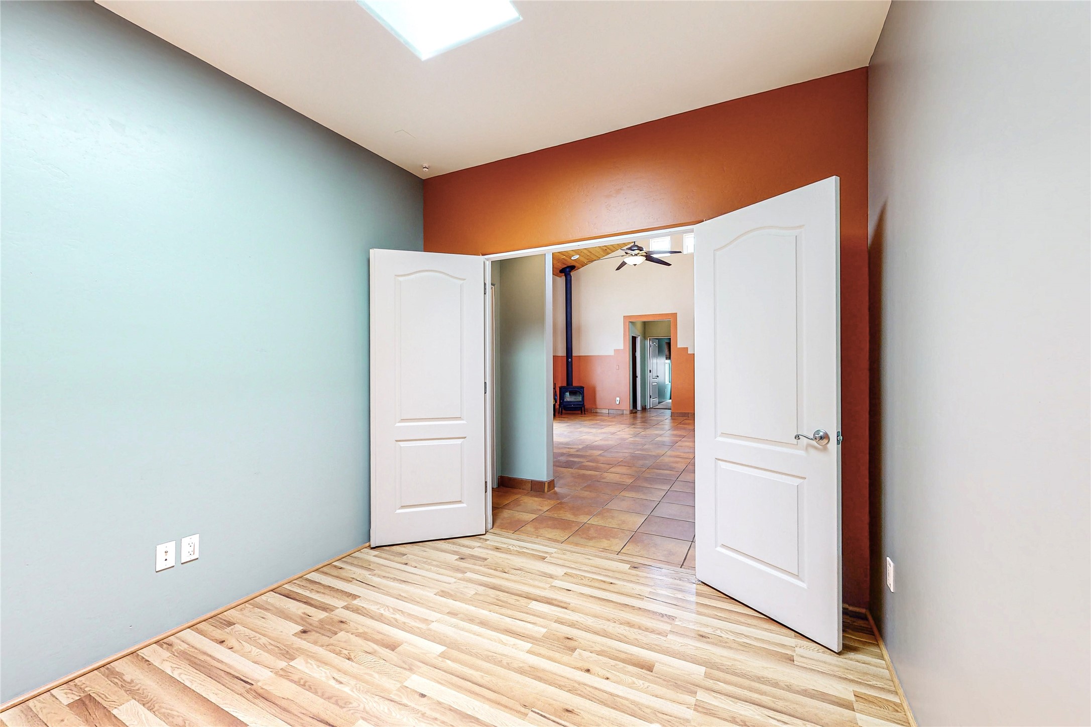Empty room with a skylight and wood-pattern flooring, looking through open doors toward a tiled room with a wood stove.