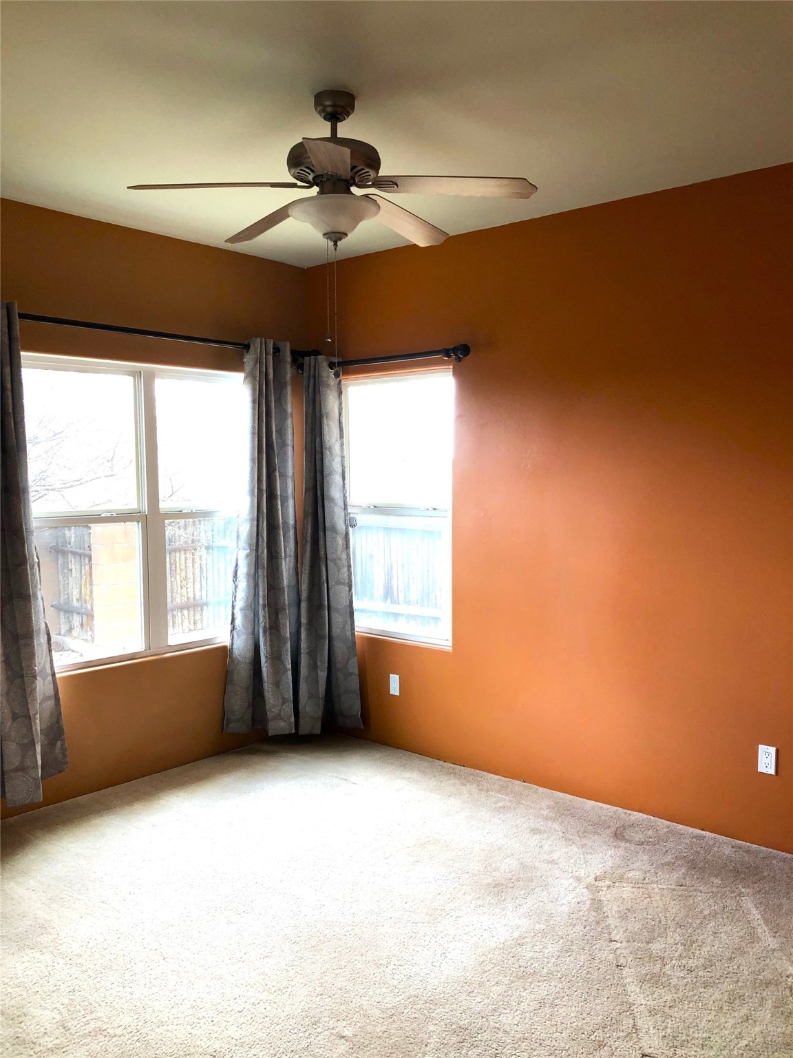 Empty bedroom with orange walls, beige carpet, two curtained windows, and a ceiling fan.