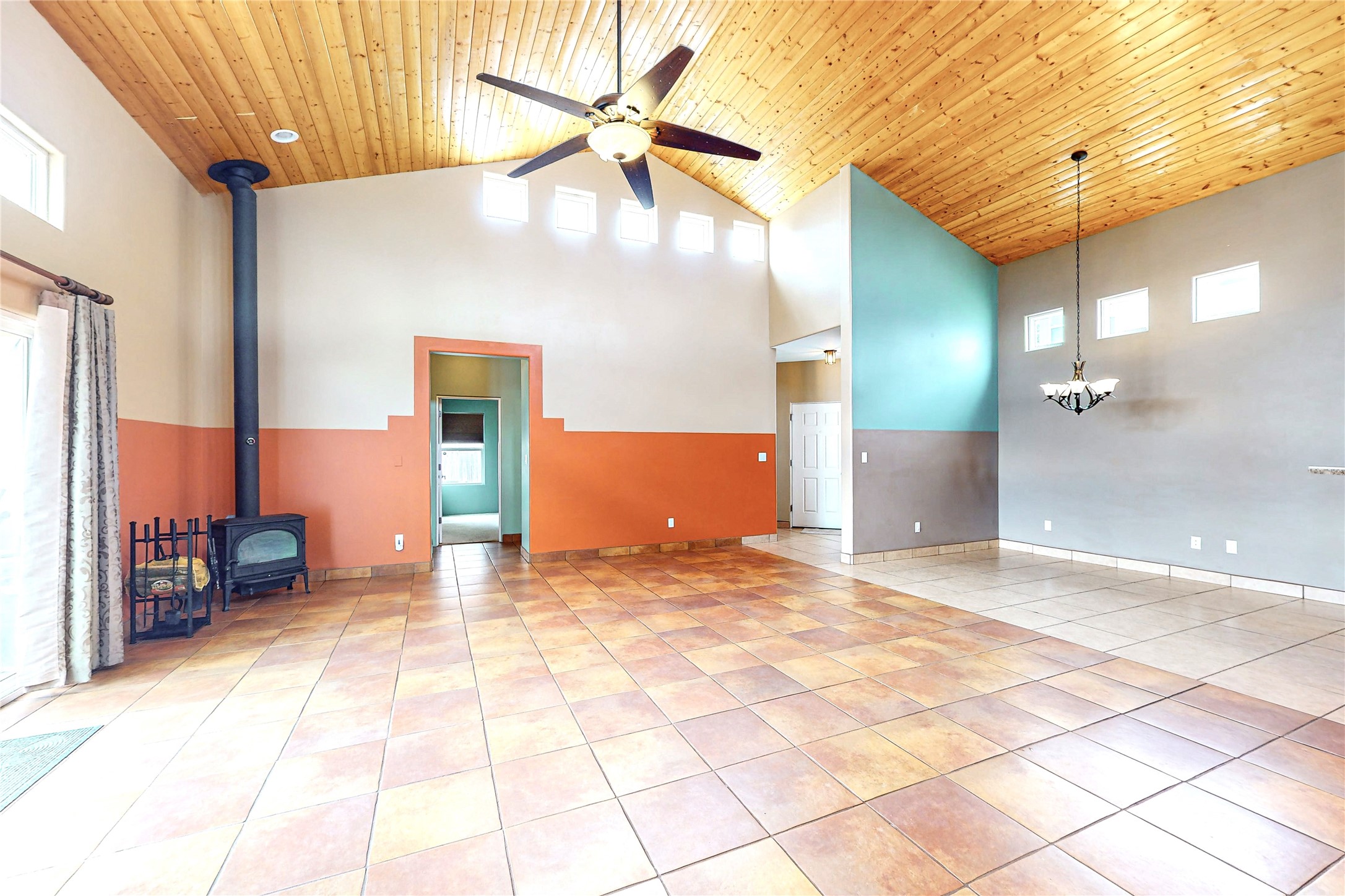 Empty living and dining area with a vaulted wood ceiling, tiled floor, and black wood stove.