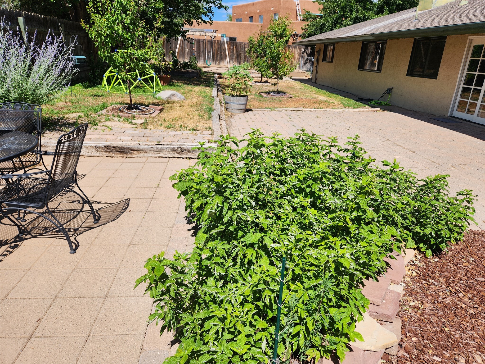 Backyard patio with black metal seating, leafy garden beds, and a small play structure.