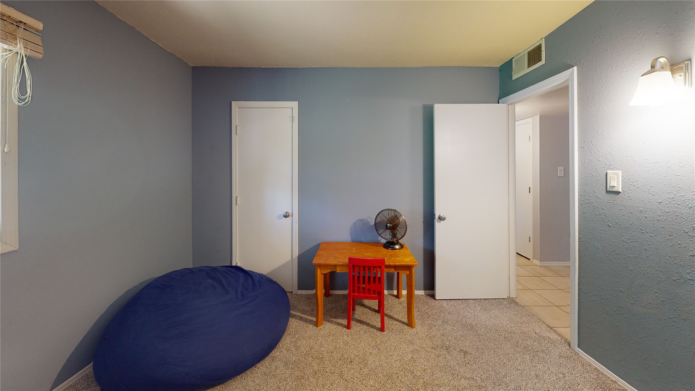 Carpeted bedroom with a navy beanbag and a wooden table with a red chair and fan.