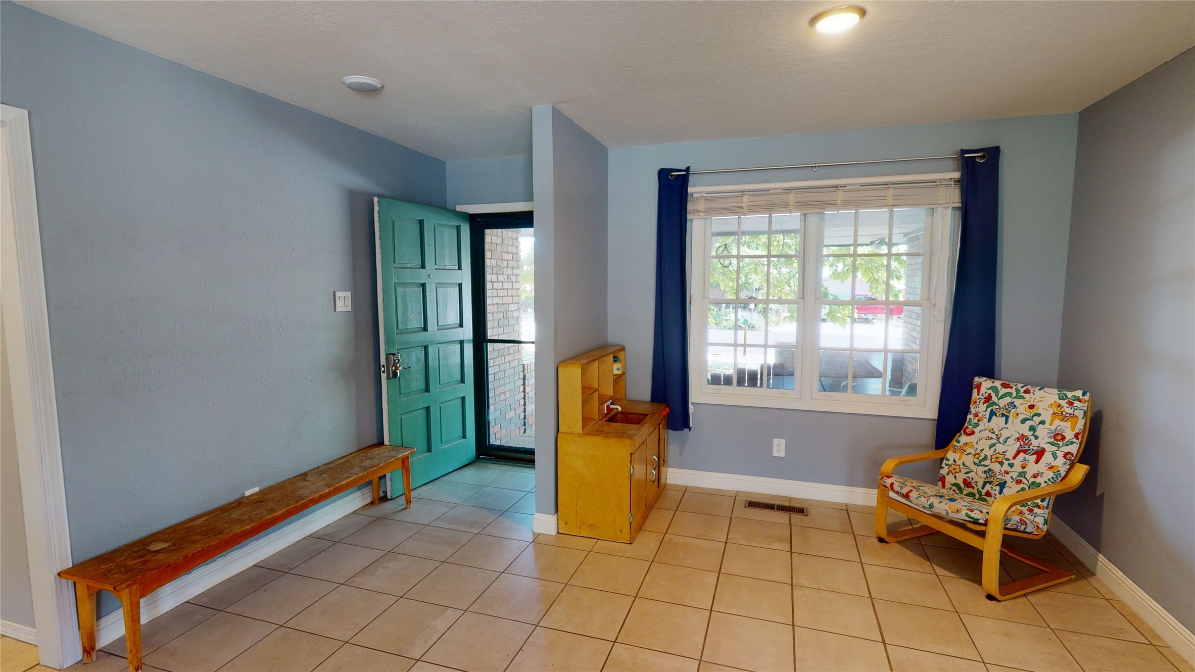 Entry room with teal front door, long wooden bench, patterned armchair, and large window.