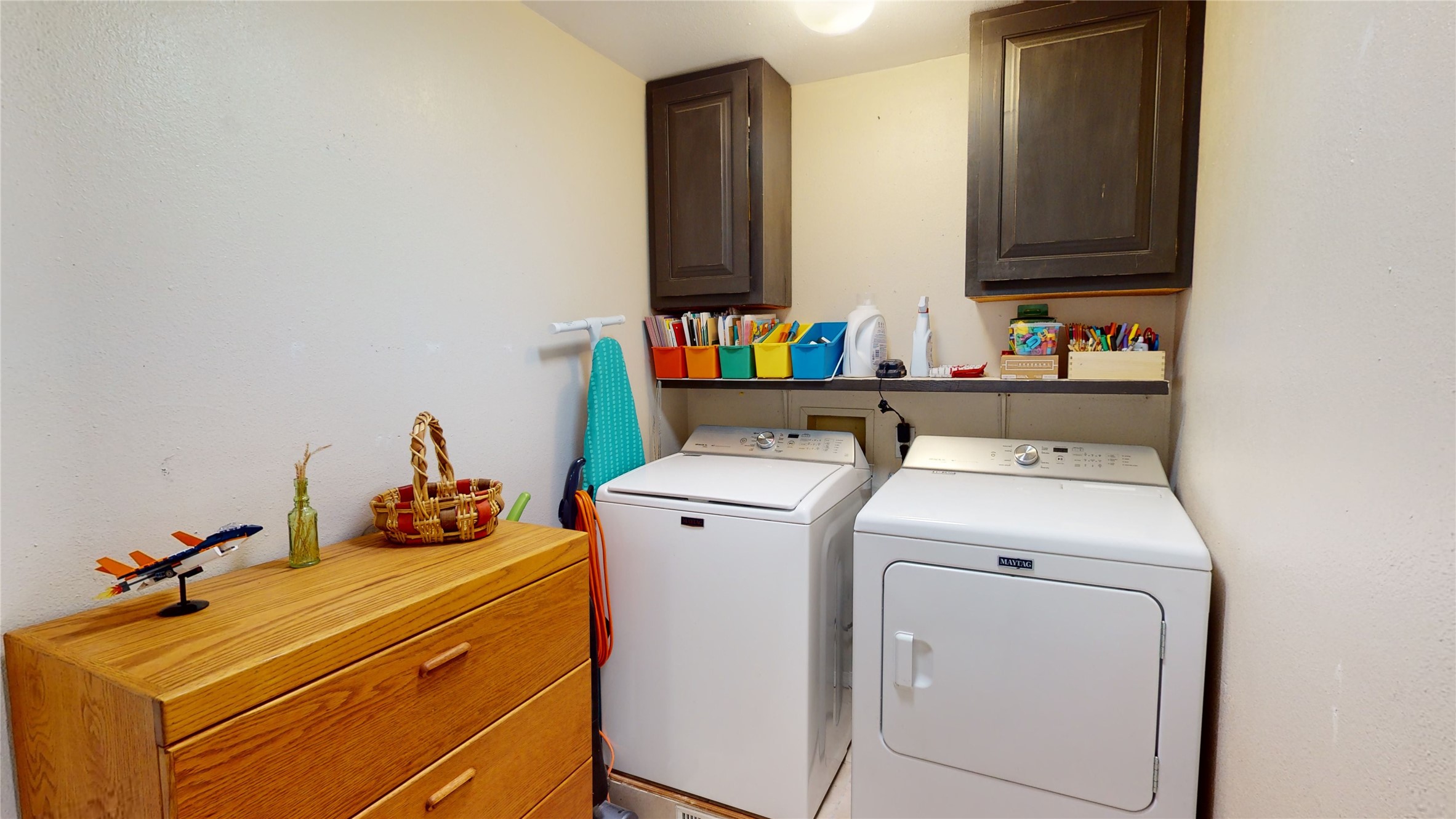 Laundry room with white washer and dryer, dark cabinets, colorful bins, and a wooden dresser.